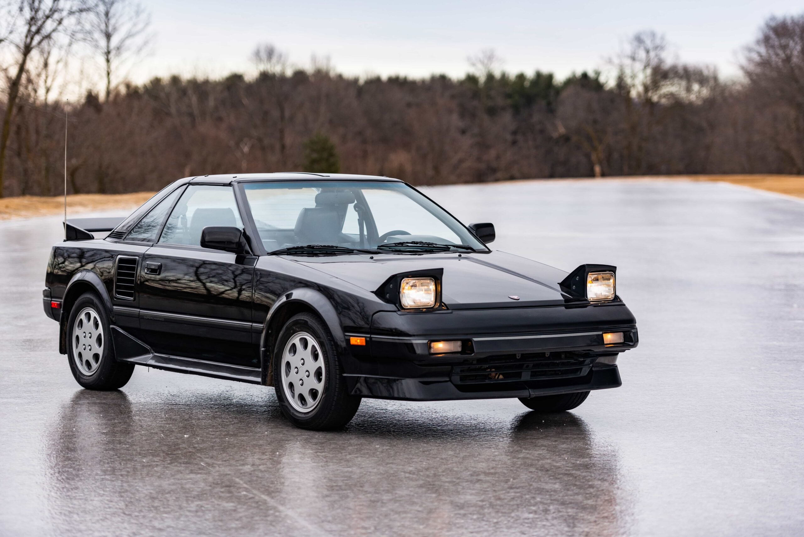 A black classic sports car with pop-up headlights is parked on a shiny surface outdoors, surrounded by trees and a cloudy sky in the background.