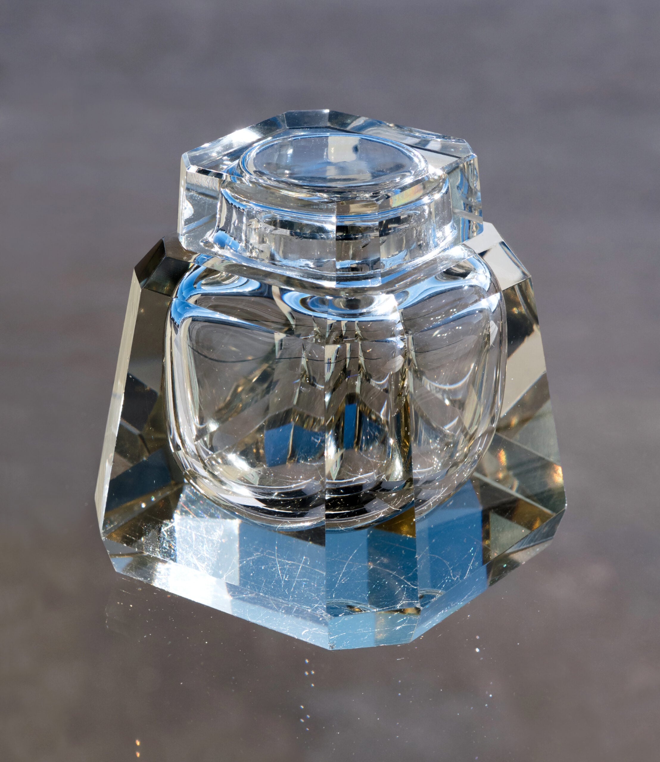 A clear, faceted glass inkwell sits on a reflective surface, casting light and blue reflections underneath.
