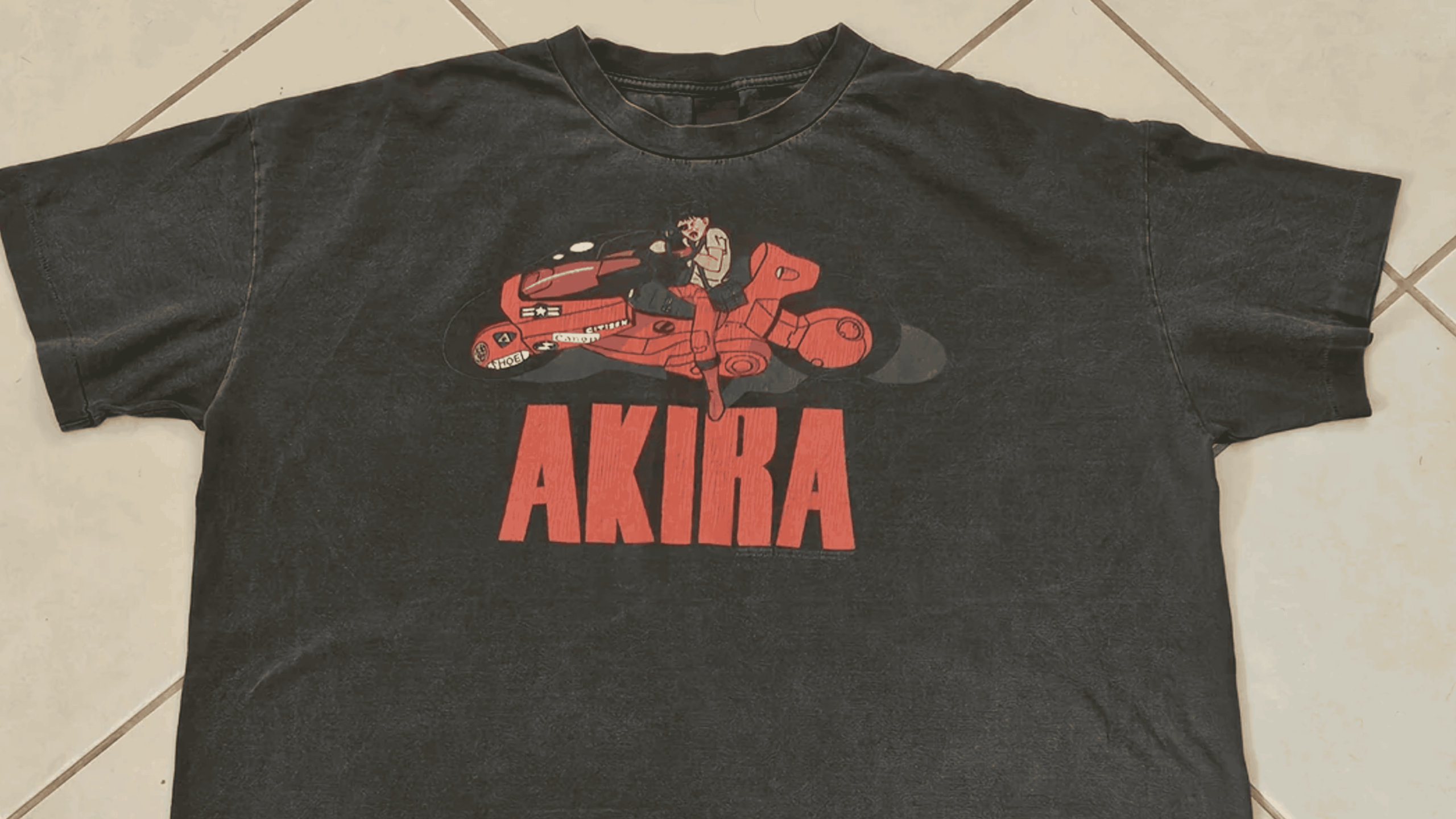 Black T-shirt featuring a graphic of a character riding a futuristic red motorcycle above bold red text that reads “AKIRA,” laid flat on tiled flooring.