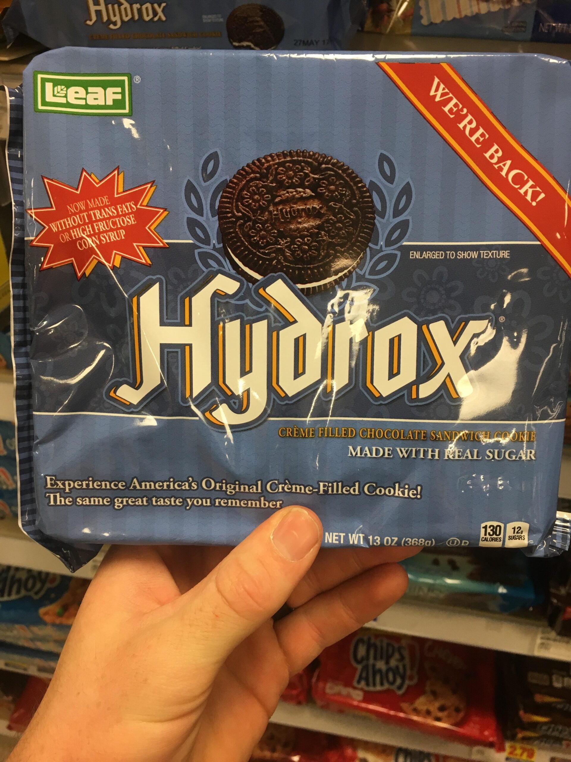 A hand holds a package of Hydrox chocolate sandwich cookies with creme filling. The package is blue and says “WE’RE BACK!” and “Made with real sugar.” Shelves with other cookies are in the background.
