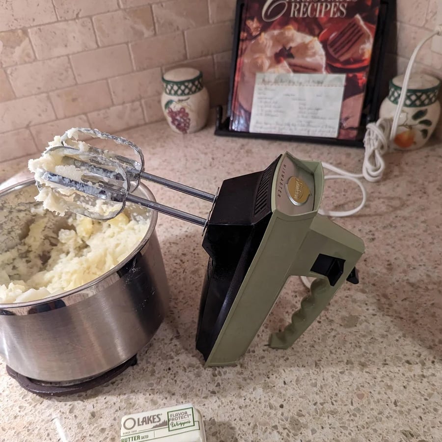 An electric hand mixer with beaters covered in mashed potatoes rests on a kitchen counter near a metal bowl, a butter stick, and a recipe book in the background.