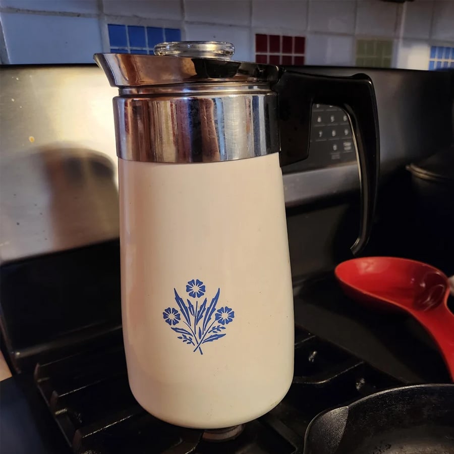 A vintage cream-colored coffee pot with a blue floral design sits on a stove, next to a red spoon. The stove’s black grates and tiled backsplash with colorful squares are visible in the background.