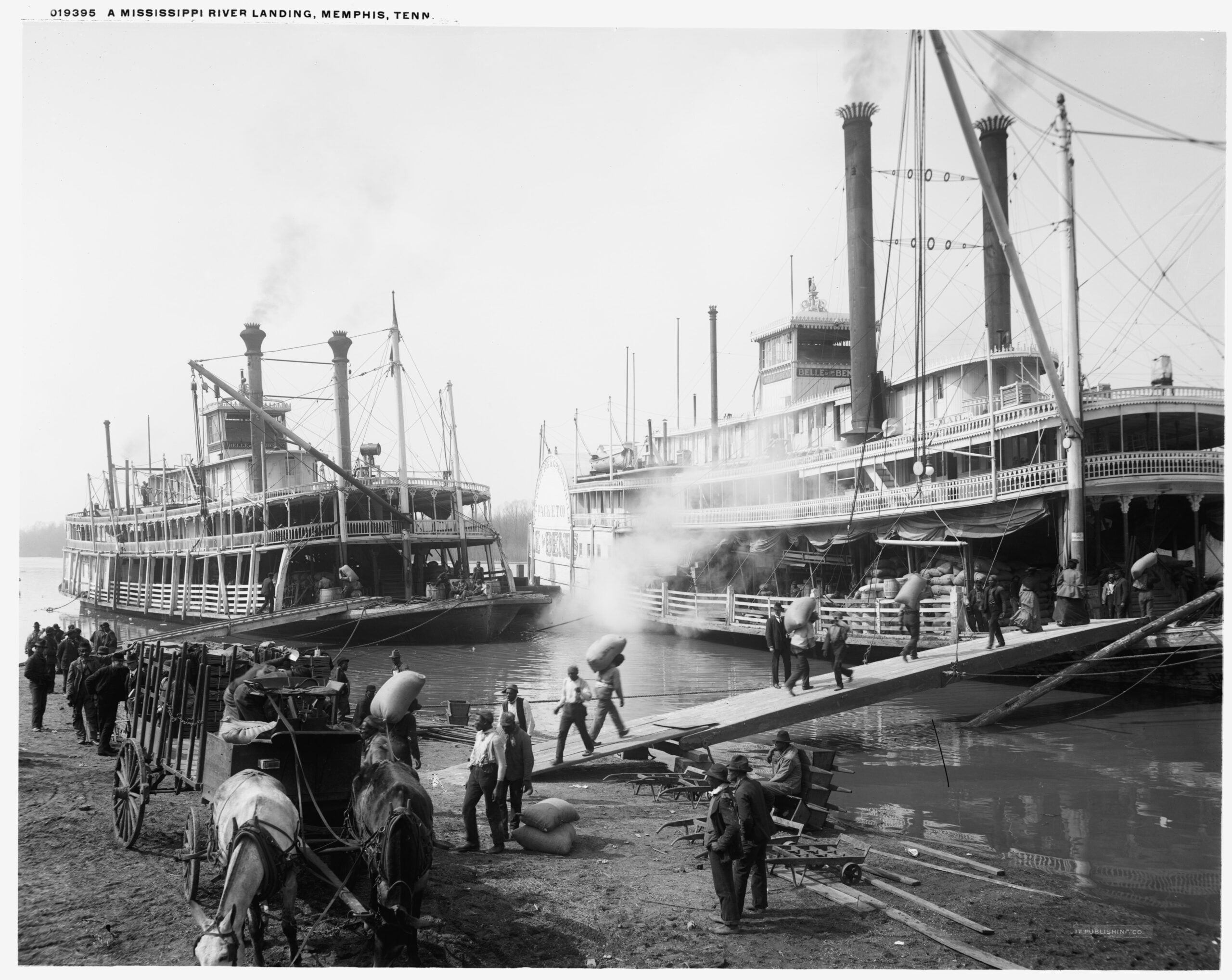 People load and unload cargo from steamboats at a busy Mississippi River landing in Memphis. Horse-drawn wagons, workers carrying sacks, and smokestacks dominate the bustling riverside scene.