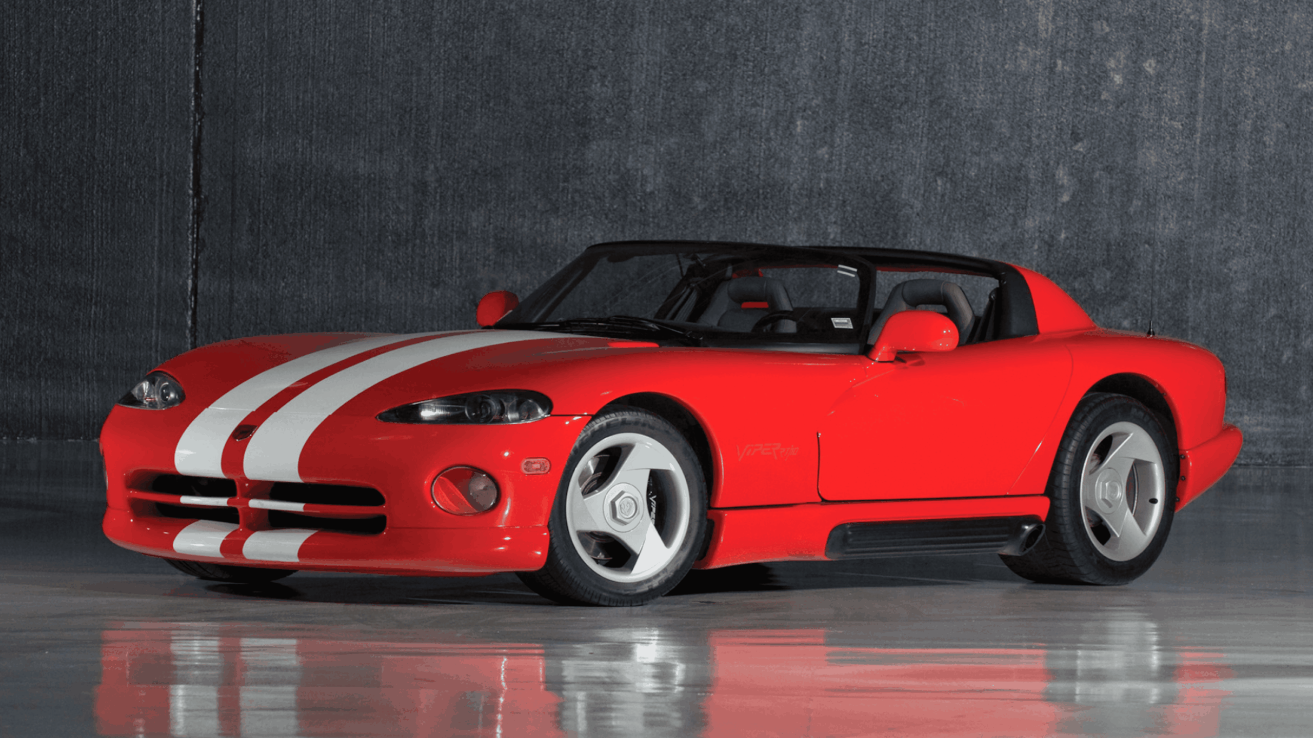A red sports car with white racing stripes, low-profile tires, and a convertible top is parked indoors on a shiny concrete floor, with a dark gray wall in the background.