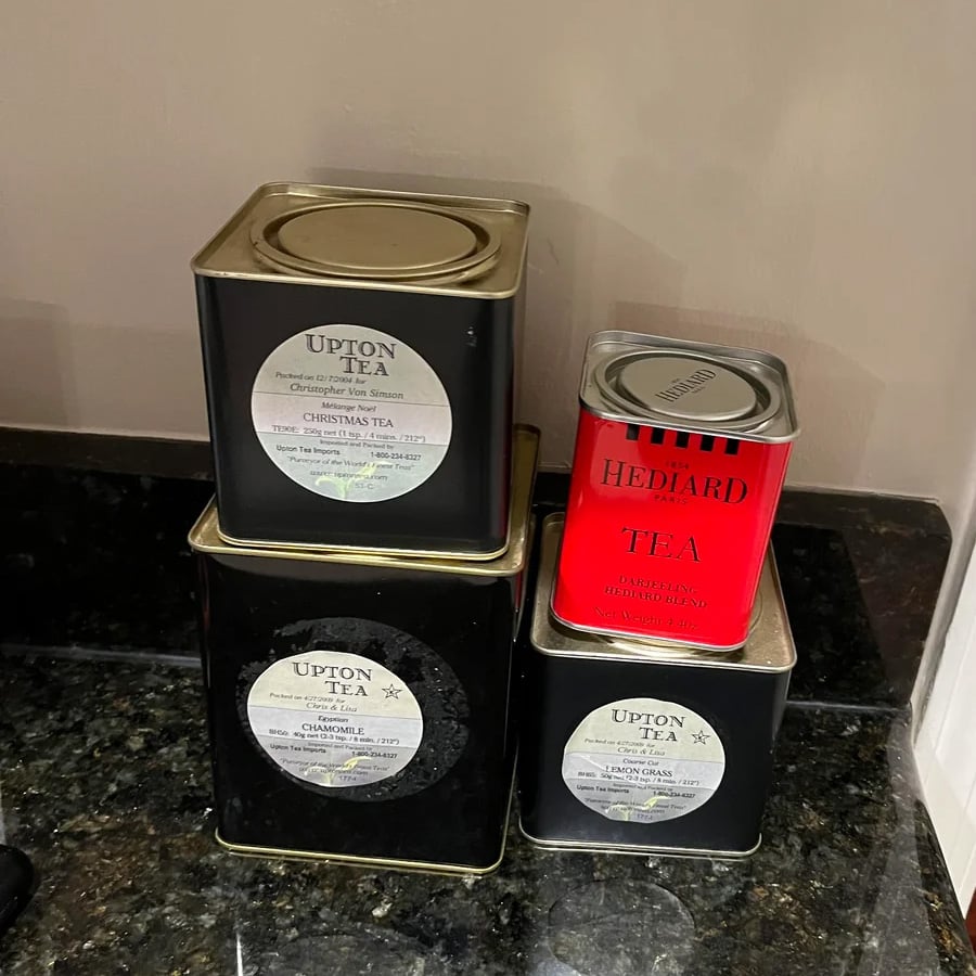 Four square tea tins are stacked on a dark countertop. Three tins are black with white labels reading “Upton Tea,” and one is smaller, red, and labeled “Hediard Tea.” All have metal lids.