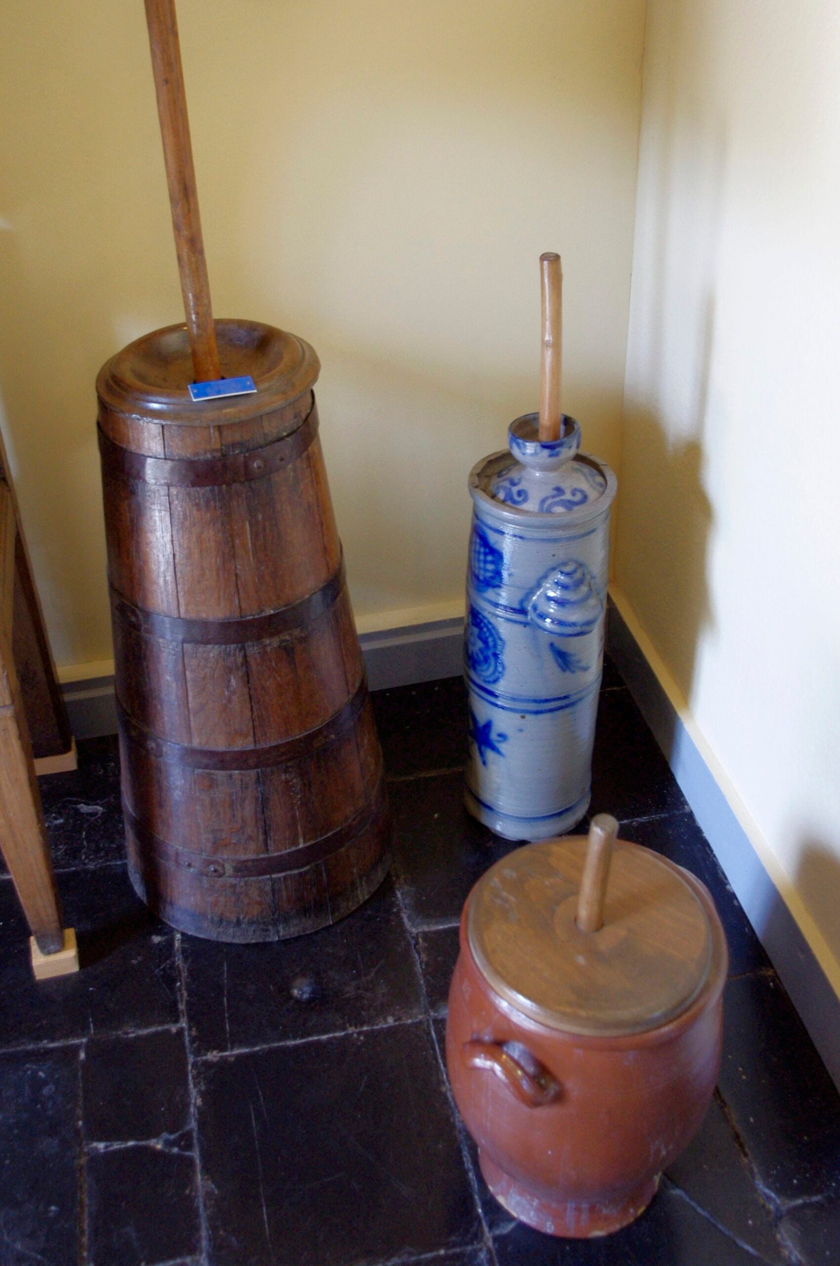 Three old-fashioned butter churns, one wooden, one blue ceramic, and one brown ceramic, stand in the corner on a dark tiled floor against a yellow wall. Each has a wooden plunger or lid.