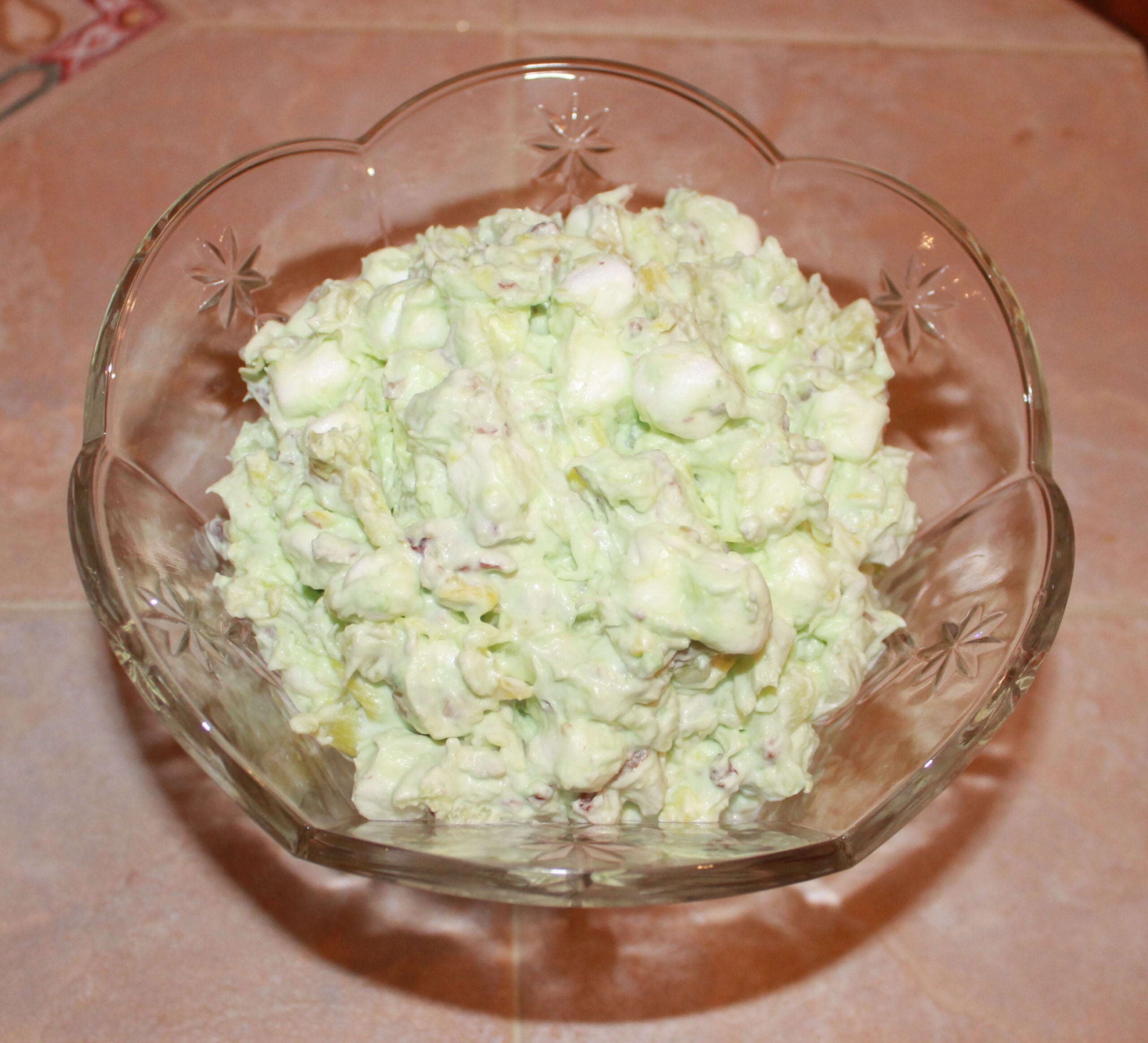 A glass bowl filled with a green-tinted creamy salad, possibly containing marshmallows, pineapple, and nuts, sits on a beige tiled surface.