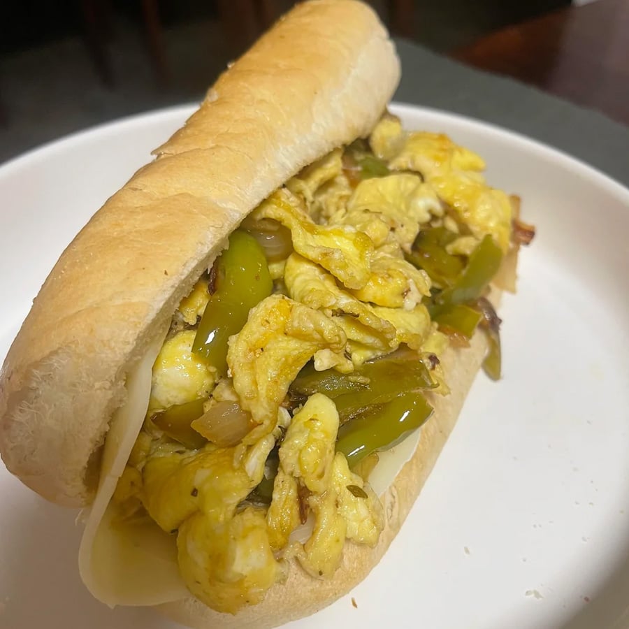 A hoagie roll filled with scrambled eggs, sautéed green bell peppers, onions, and melted cheese, served on a white plate.
