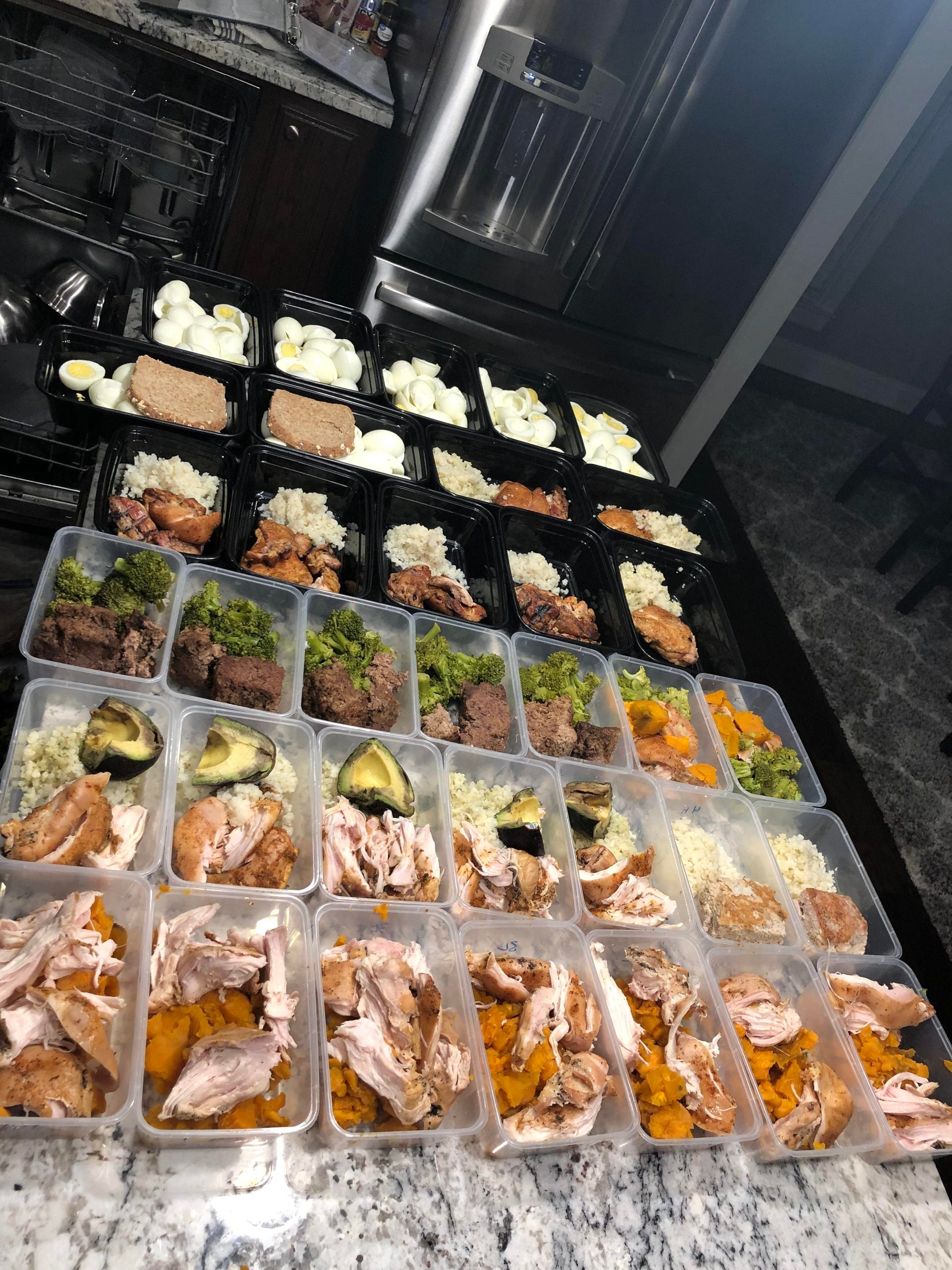 A kitchen counter with many meal prep containers filled with rice, chicken, broccoli, eggs, sweet potatoes, and avocado. Hard-boiled eggs and bread are also visible near a dishwasher and refrigerator.