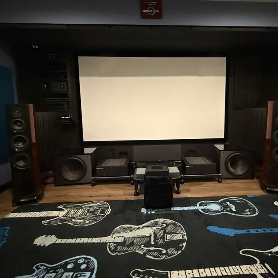 A home theater room with a large projector screen, multiple speakers, AV equipment, and a rug featuring guitar designs.
