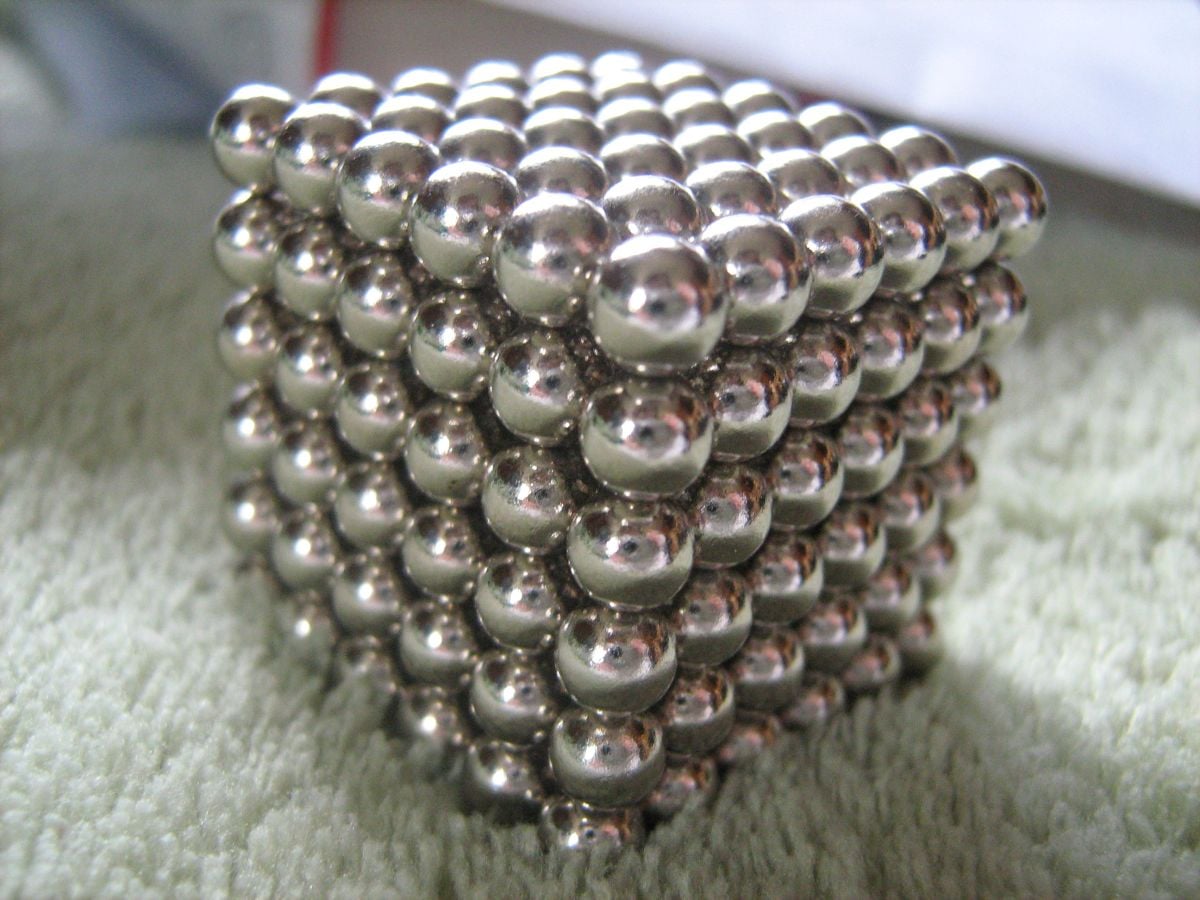 A cube made of small, shiny silver magnetic balls is arranged neatly on a textured, light green surface.
