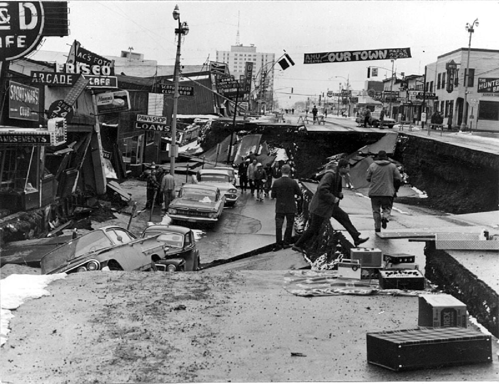 People observe a massive street collapse in a city, with cars stranded and buildings damaged. Tilted signs and debris are visible, while bystanders and officials examine the scene under cloudy skies.