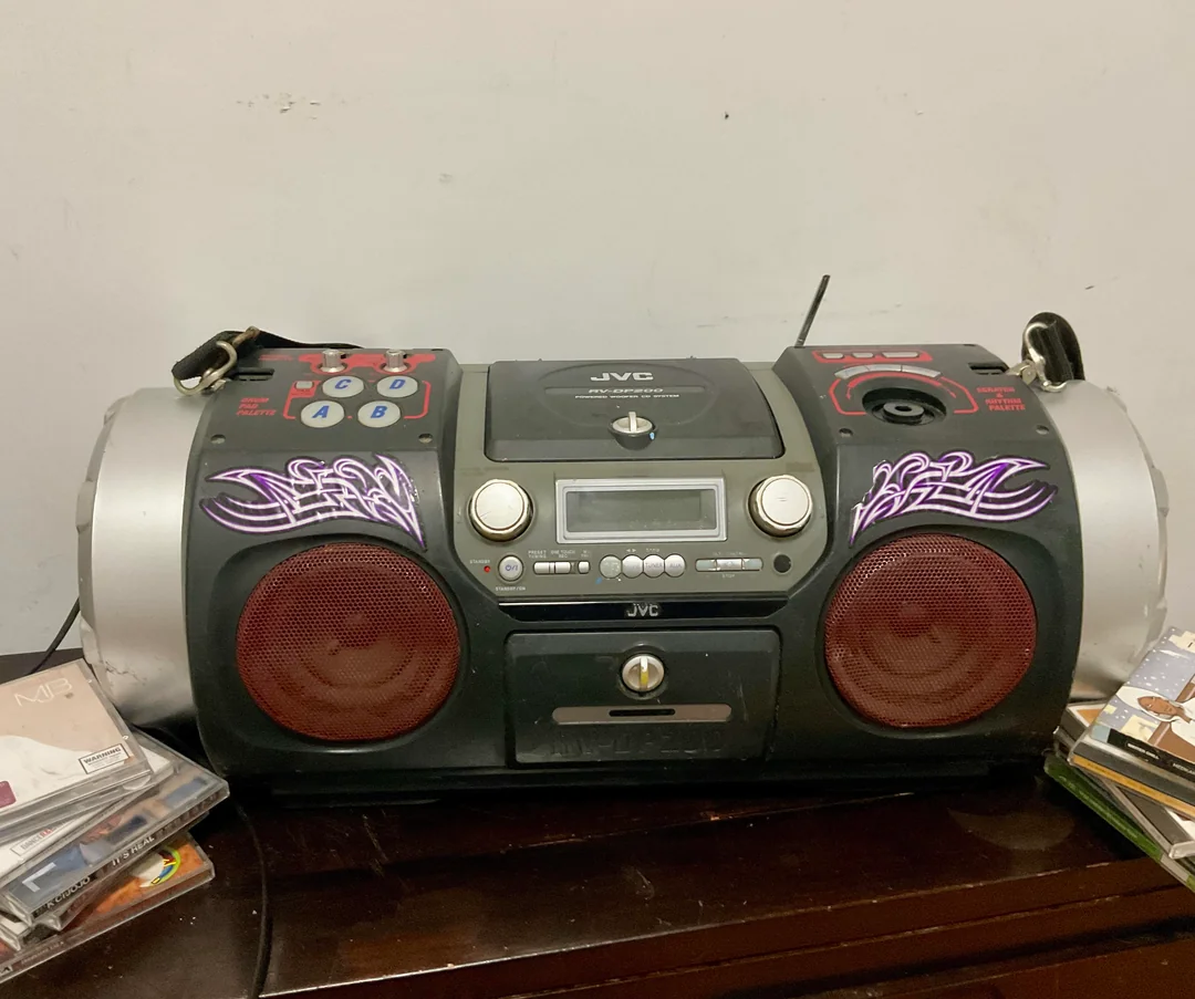 A black and silver JVC boombox with red speakers, graffiti-style purple decals, and various CDs stacked nearby, sits on a dark wooden surface against a plain white wall.