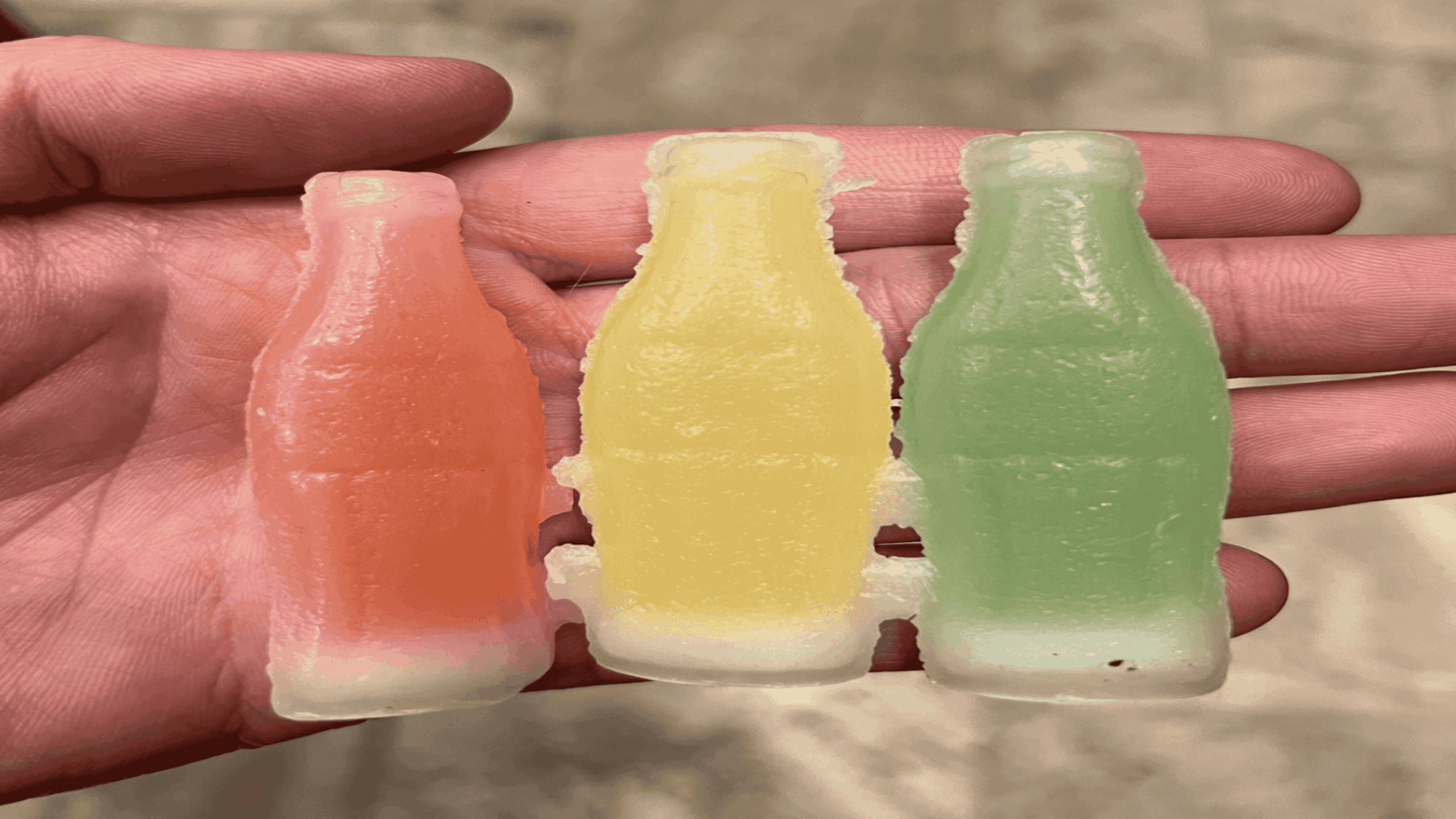 A hand holds three gummy candies shaped like soda bottles. The candies are colored red, yellow, and green, each with a white base, and are detailed to resemble classic glass soda bottles.