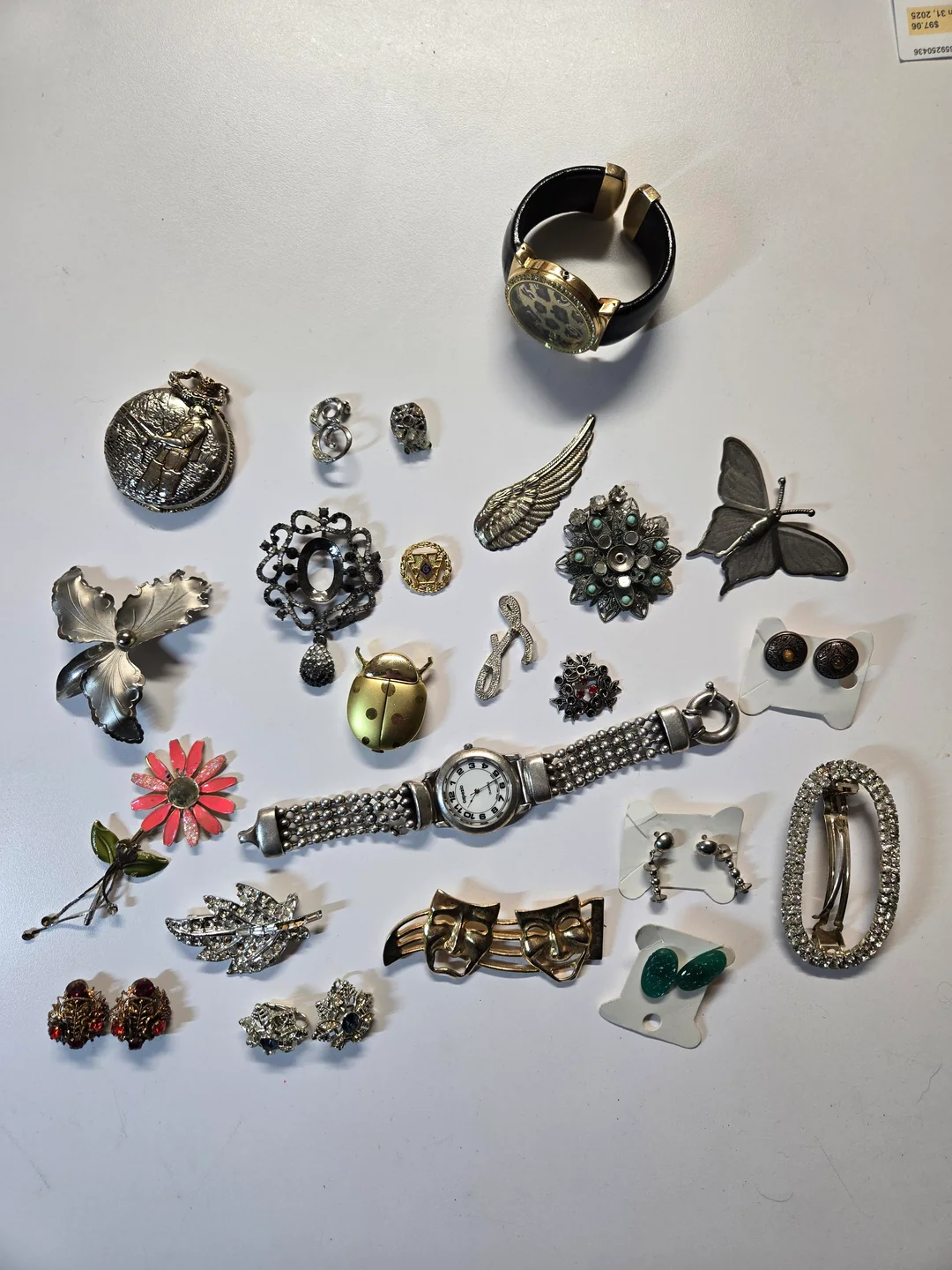A collection of jewelry and accessories, including brooches, earrings, watches, pins, and a bracelet, arranged on a white surface. Items feature floral, insect, and vintage designs in various metallic tones.