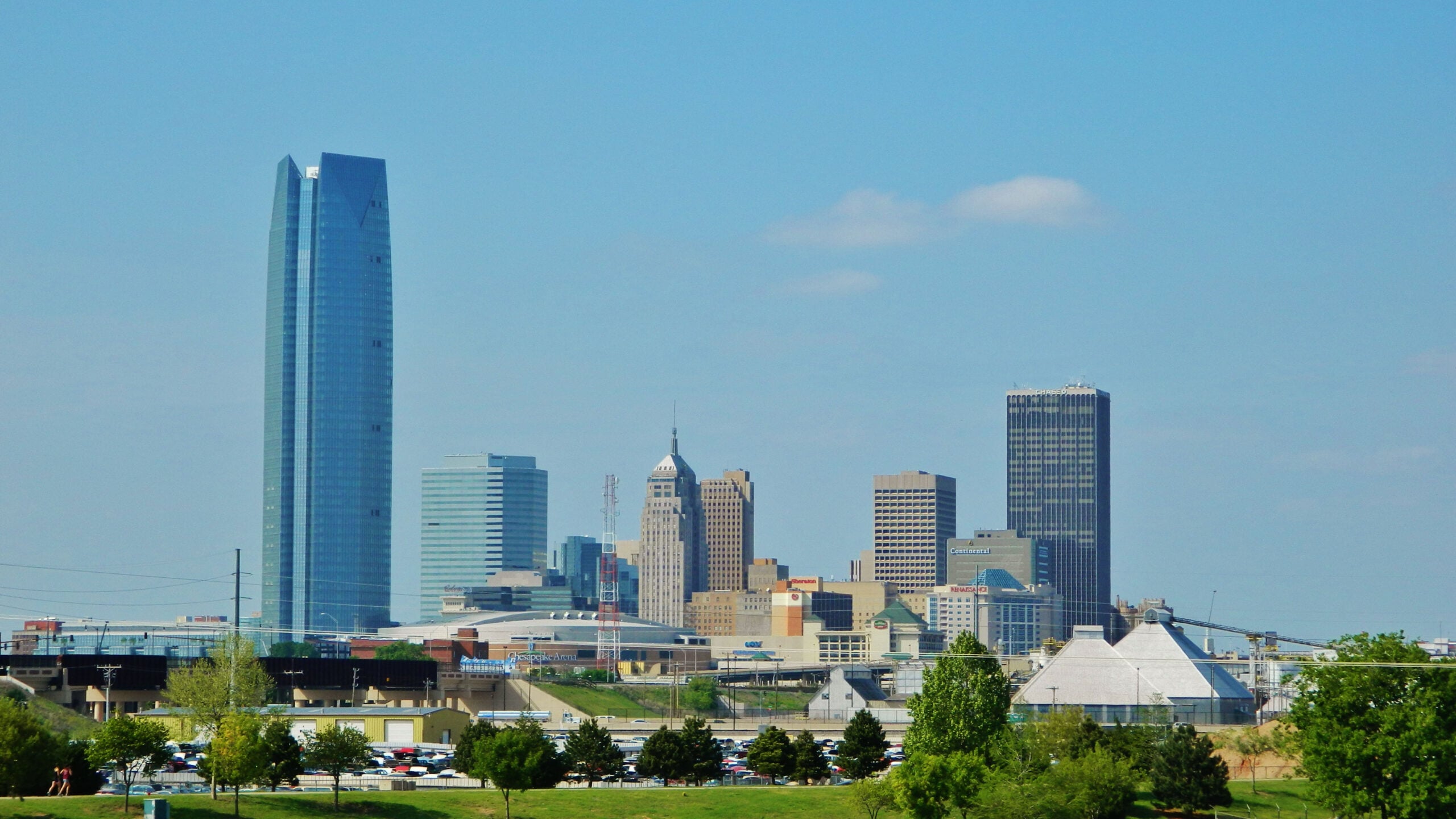 Oklahoma City skyline on a clear day, featuring tall skyscrapers, green trees in the foreground, and a blue sky with a few clouds overhead.
