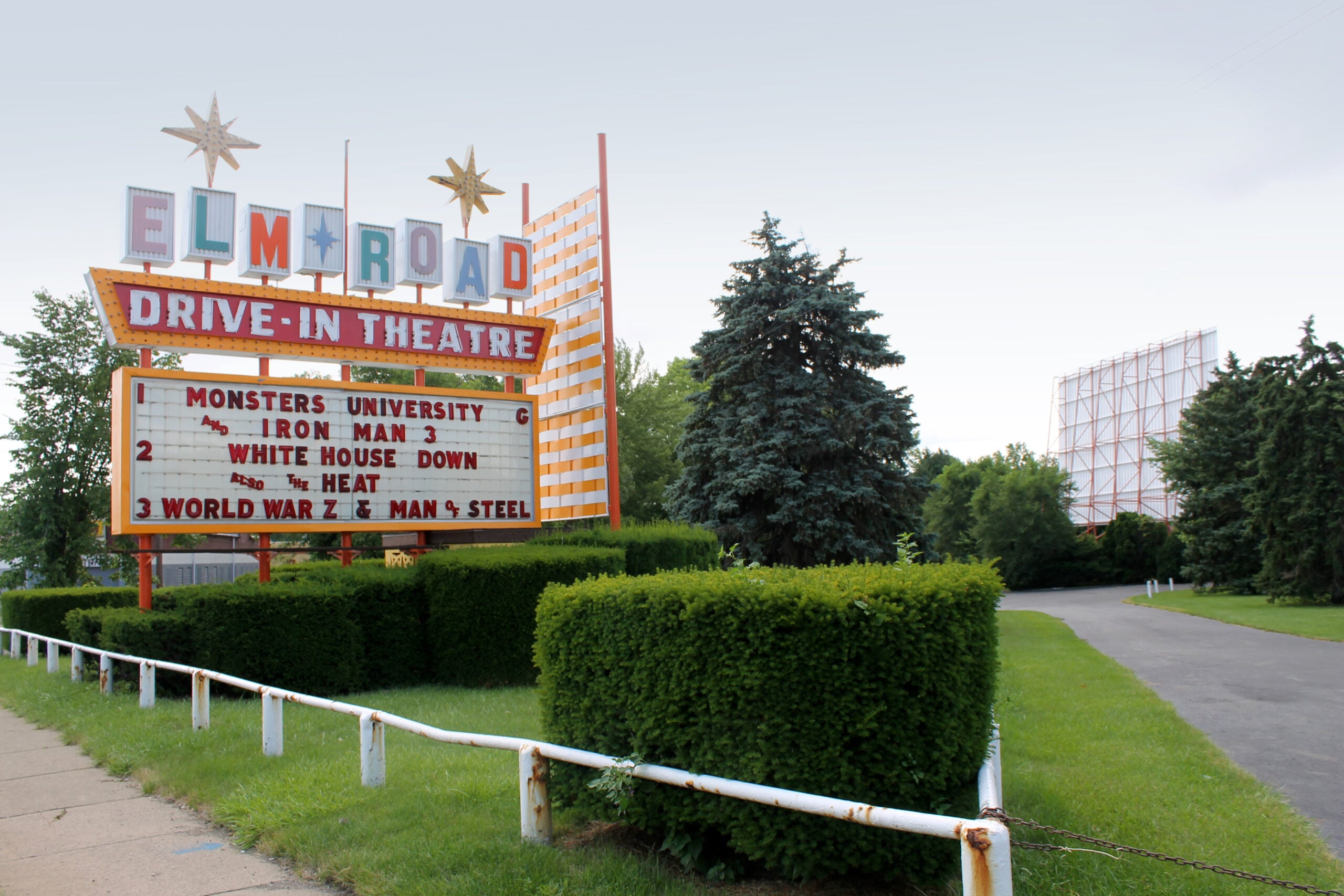 ELM ROAD DRIVE-IN THEATRE sign lists movies on its marquee, surrounded by neatly trimmed hedges and trees, with movie screens visible in the background under a cloudy sky.