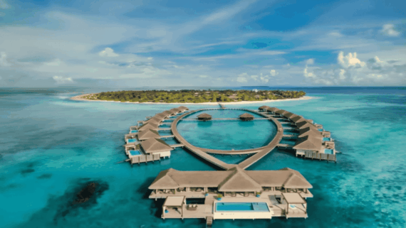 Overwater villas form a teardrop shape above turquoise ocean waters, with a lush green island and sandy beach in the background under a blue sky with scattered clouds.