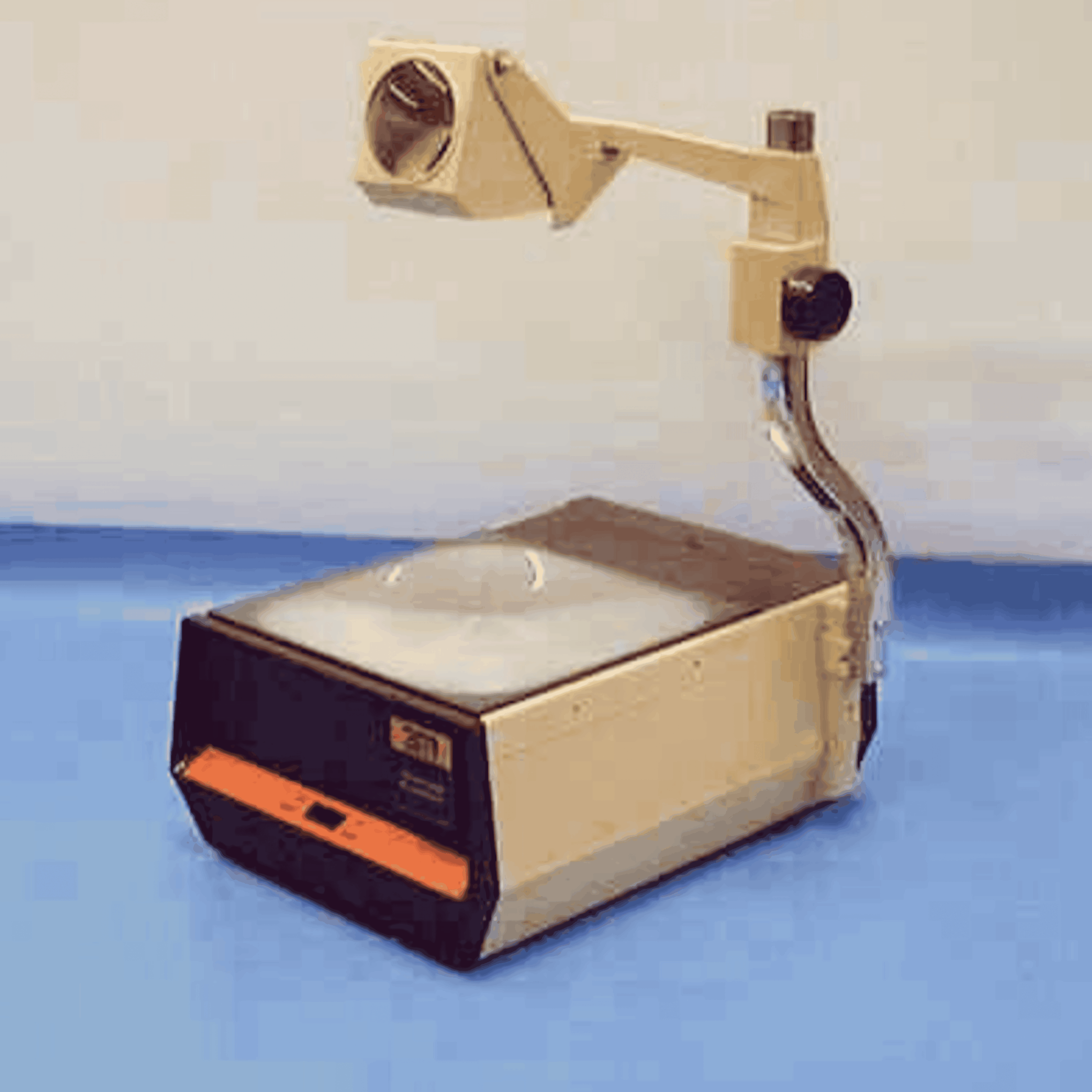 An overhead projector with a glass stage, mirror, and lens arm sits on a blue surface against a light background.