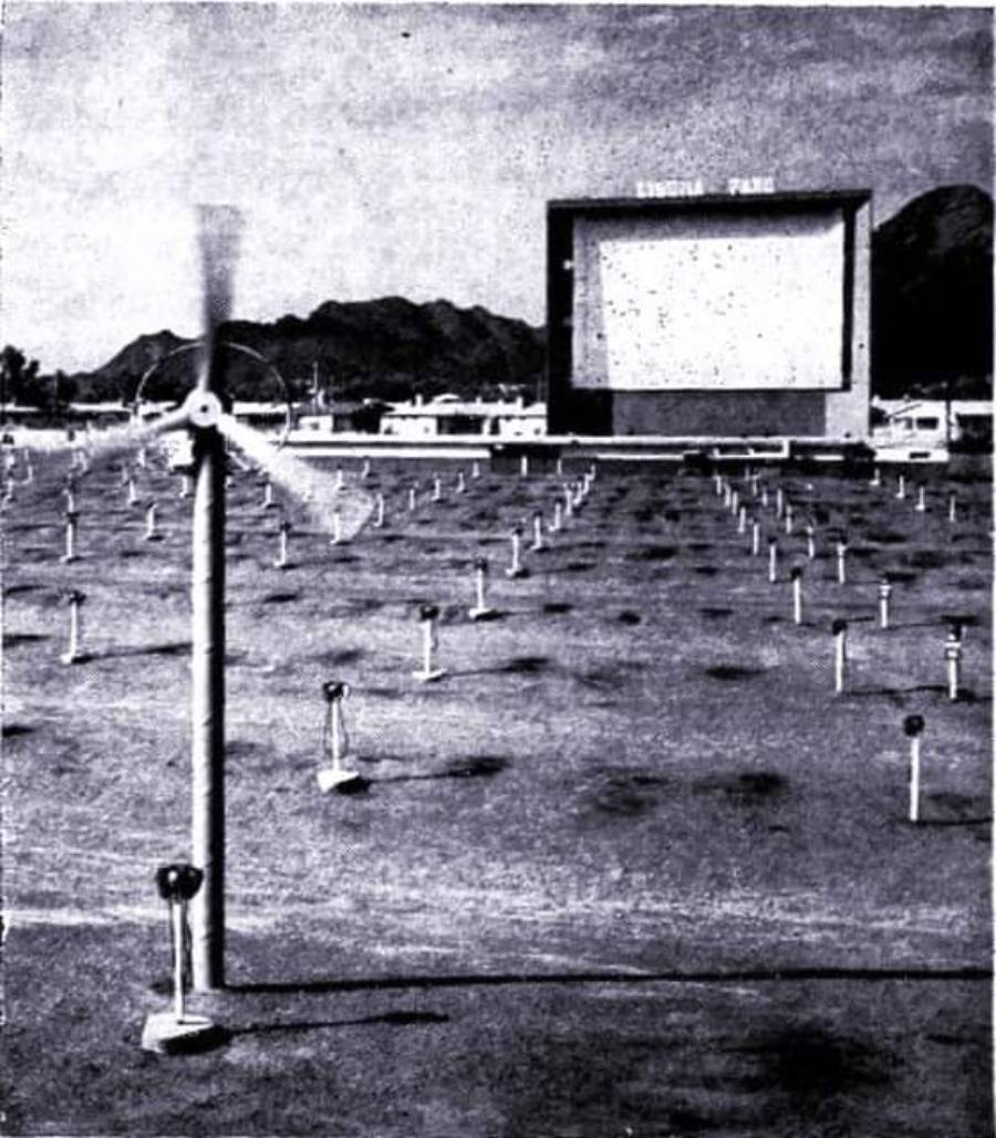 Black-and-white photo of a drive-in theater with rows of parking spots and speaker stands, a large movie screen in the background, and a spinning windmill in the foreground. Mountains are visible in the distance.