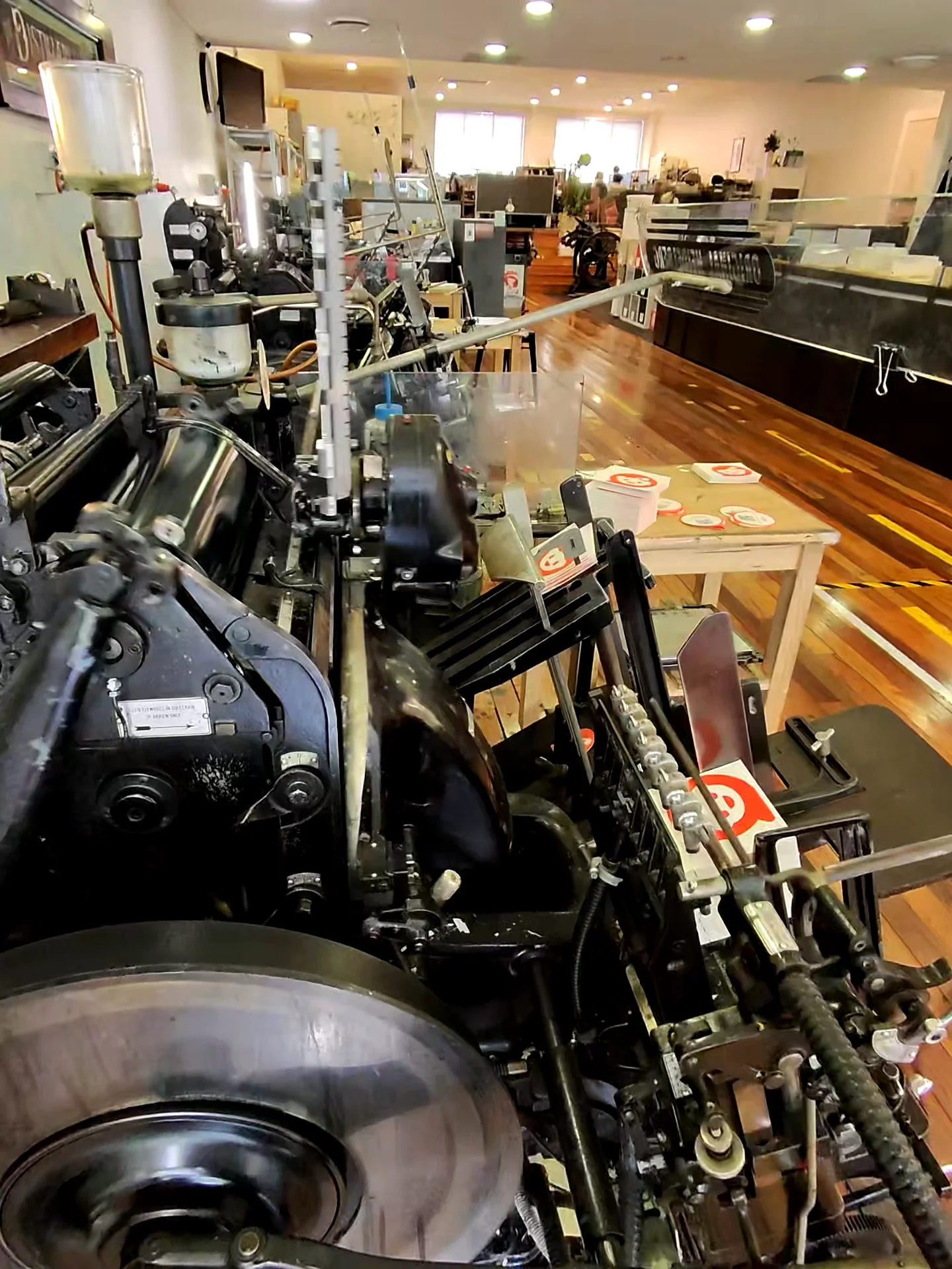 Close-up of an old printing press in a well-lit room with wooden floors, other machines, and tables displaying printed materials in the background.
