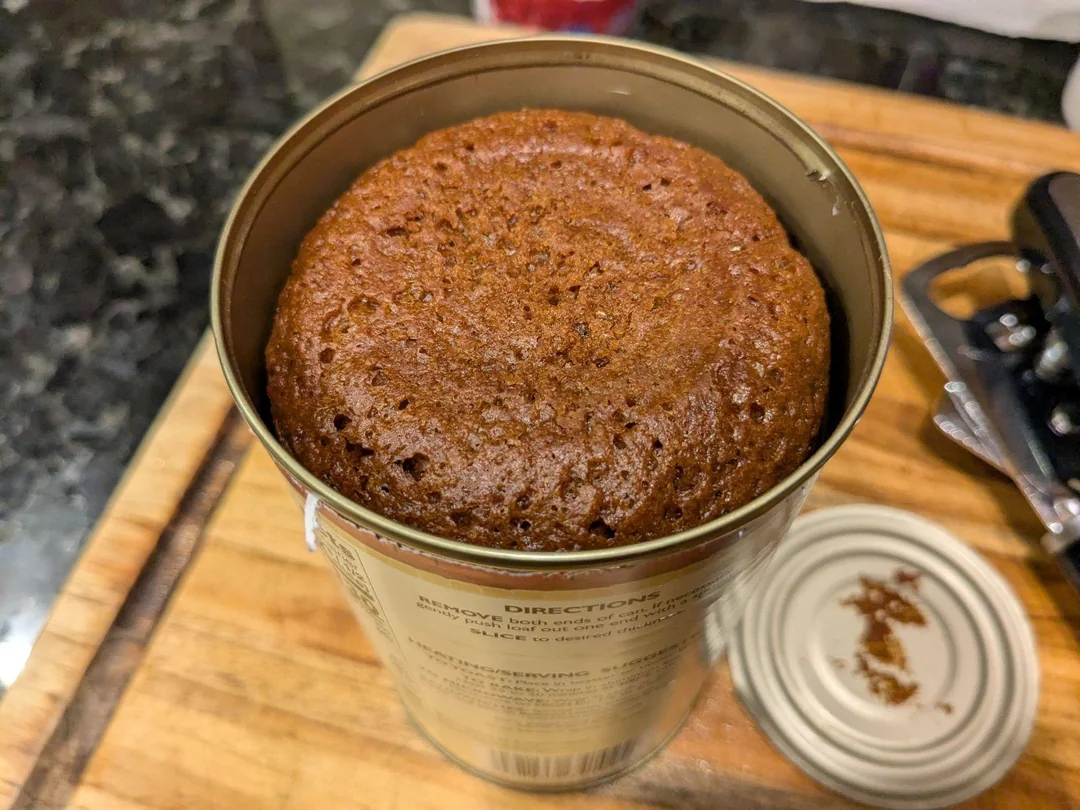 A loaf of brown bread is baked inside an opened can, sitting on a wooden cutting board with the lid removed and a can opener nearby.