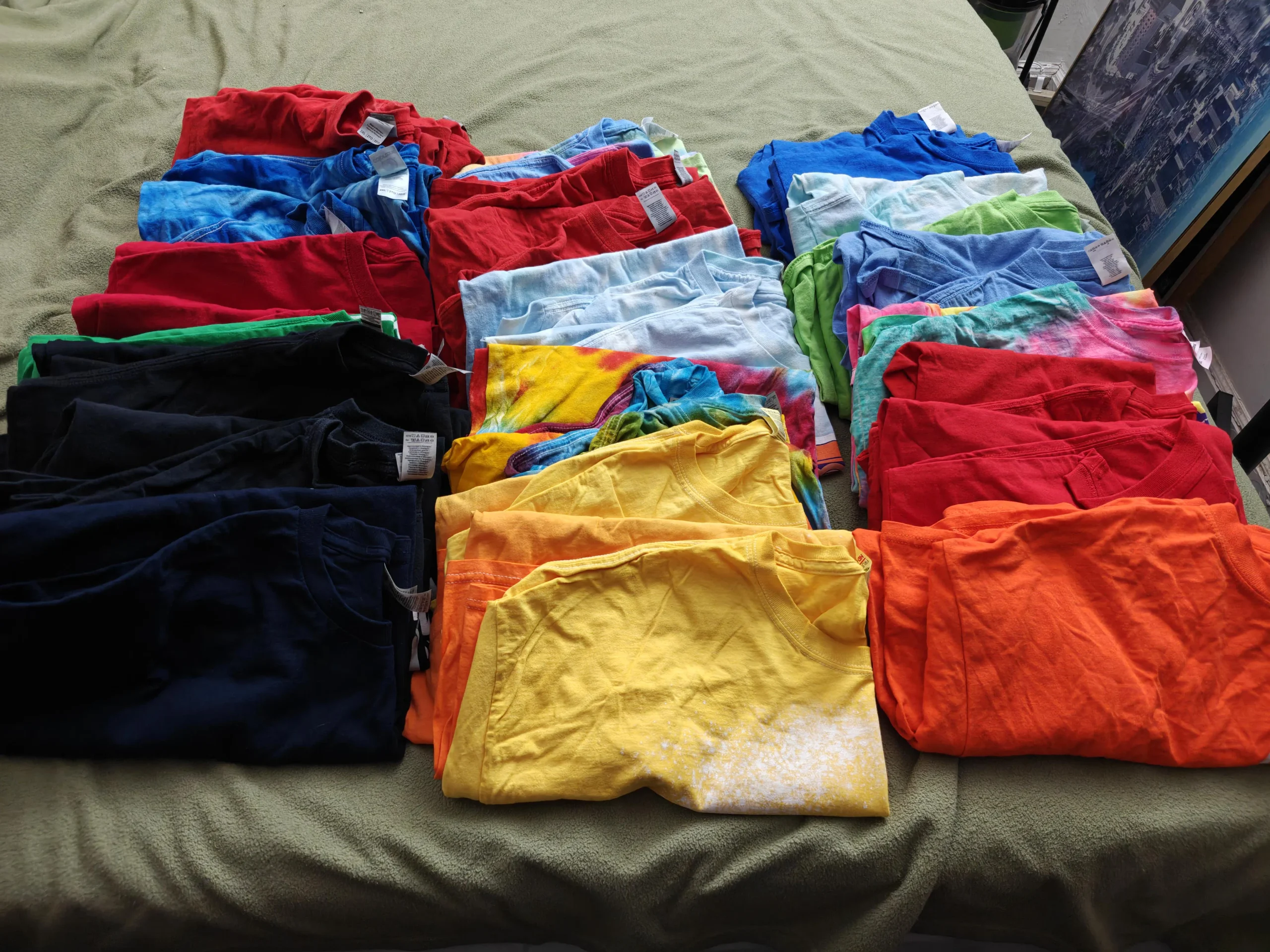 Neatly folded t-shirts in various colors, including blue, red, green, black, yellow, and orange, are arranged in rows on a bed with a green blanket.