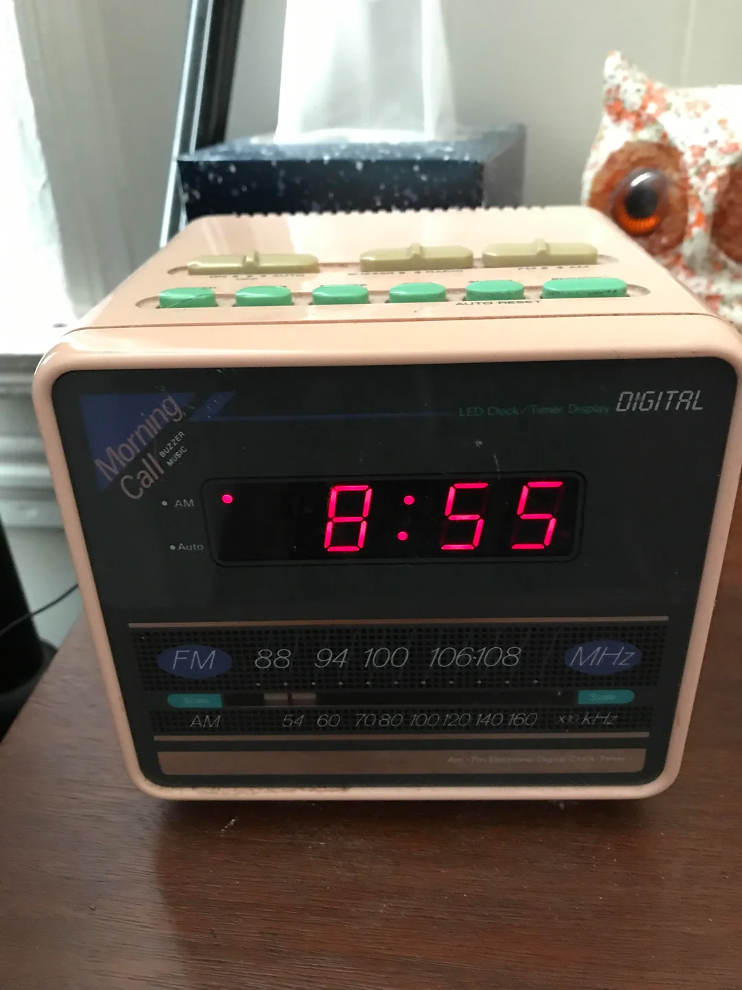 A digital clock radio displays the time 8:55 in red numbers. It has buttons on top and a frequency display on the front. An owl-shaped object and window are in the background.