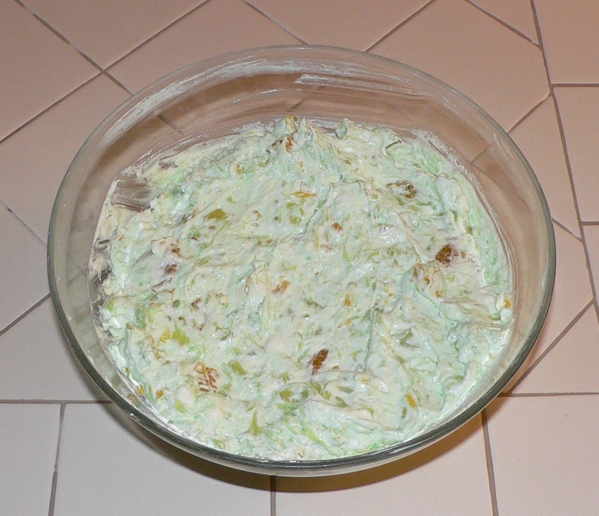 A glass bowl filled with light green, creamy dessert salad, possibly containing chunks of fruit, sits on a tiled countertop.