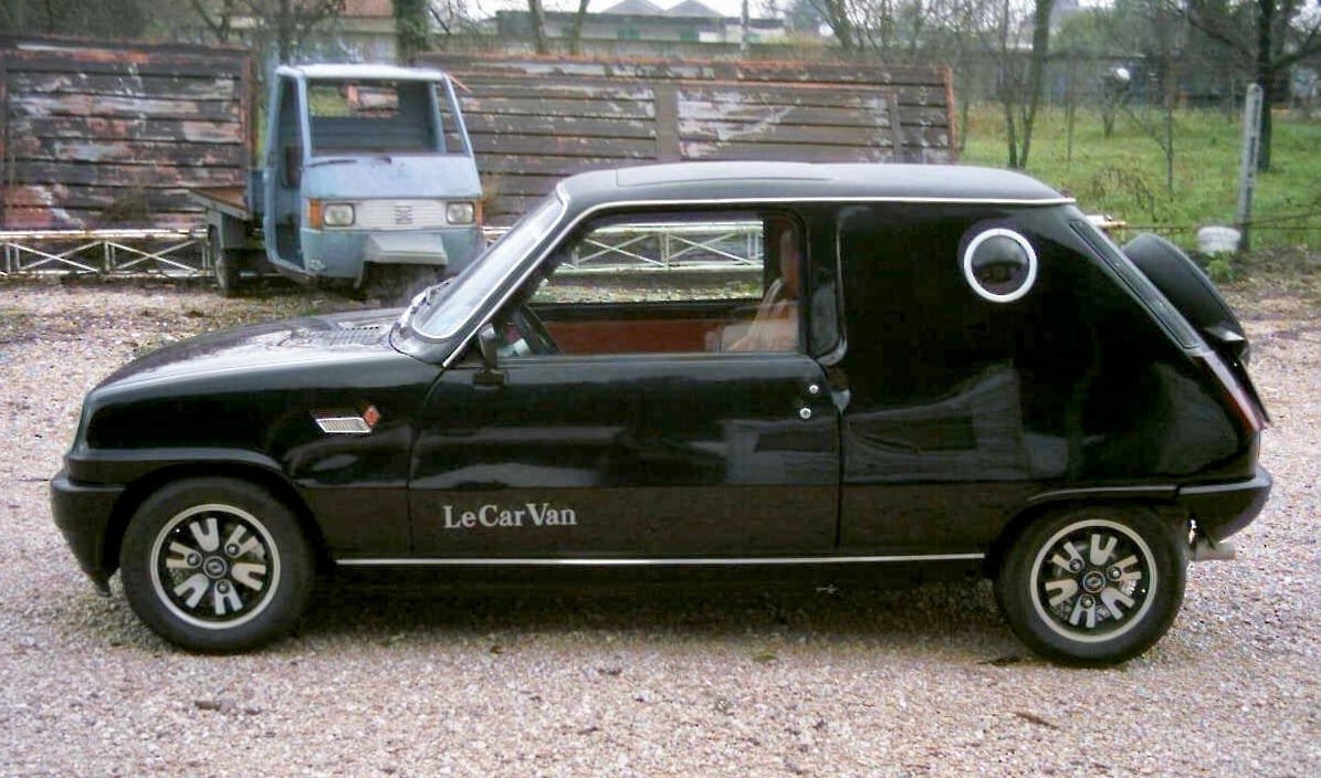 A small, black vintage car labeled “Le CarVan” is parked on gravel, with a rusty three-wheeled vehicle and wooden fence in the background.
