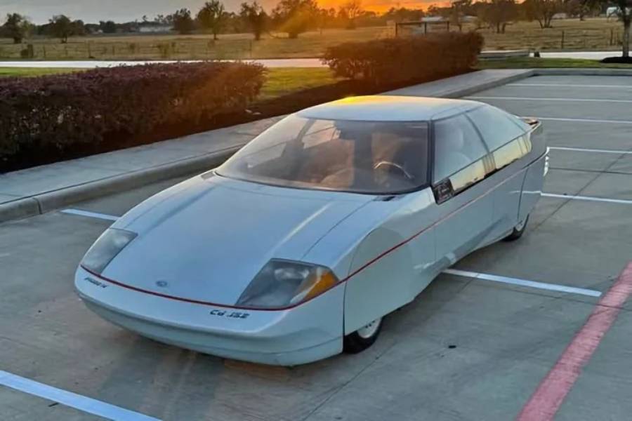 A futuristic, low-profile silver car with a sleek, aerodynamic design is parked in an empty lot at sunset. The car has a unique, smooth body and covered rear wheels.