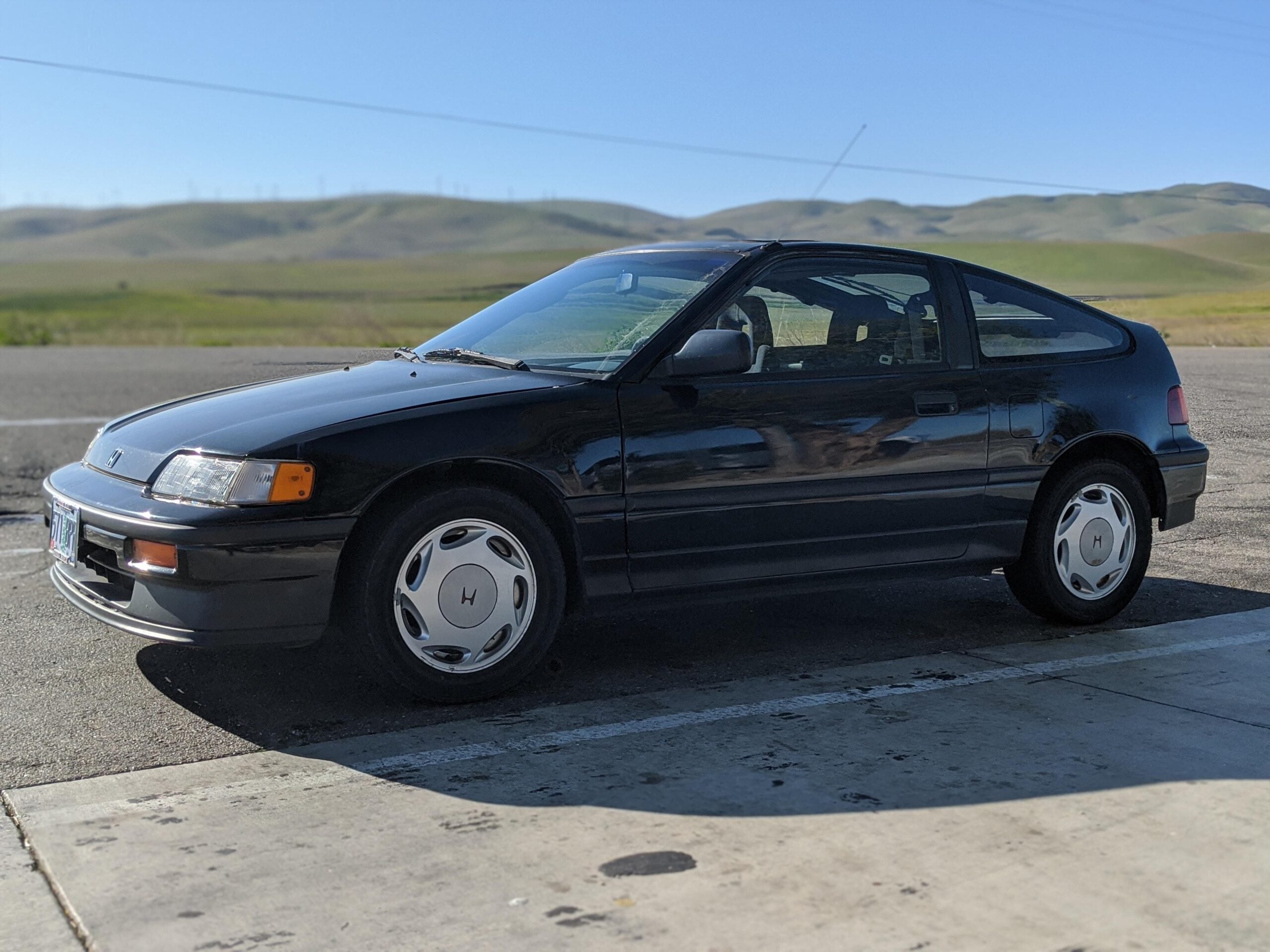 A black two-door Honda CRX hatchback is parked on concrete near an open field with hills in the background under a clear blue sky.