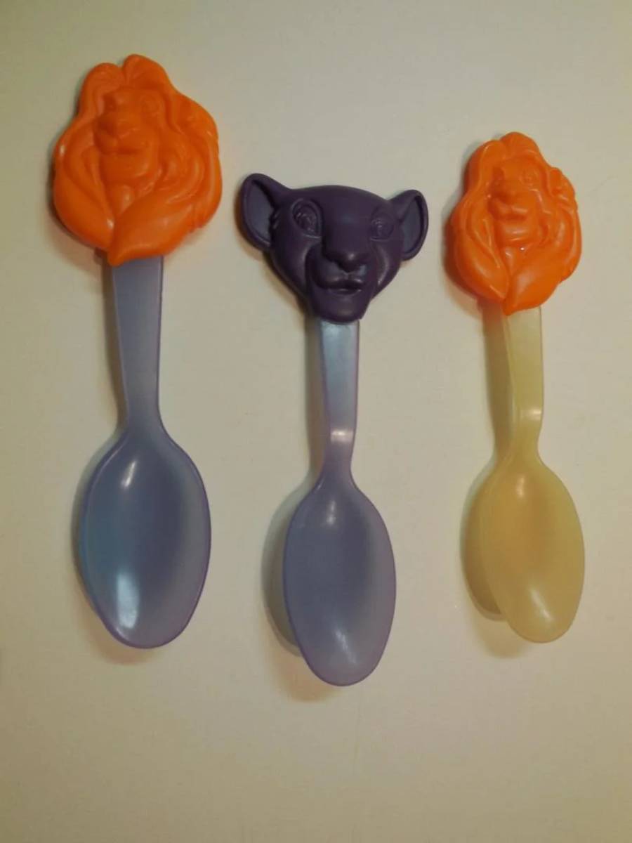 Three plastic spoons with Lion King character faces on the handles: two orange lion faces on the left and right, and a purple lion face in the center. The spoon bowls are blue and yellow.