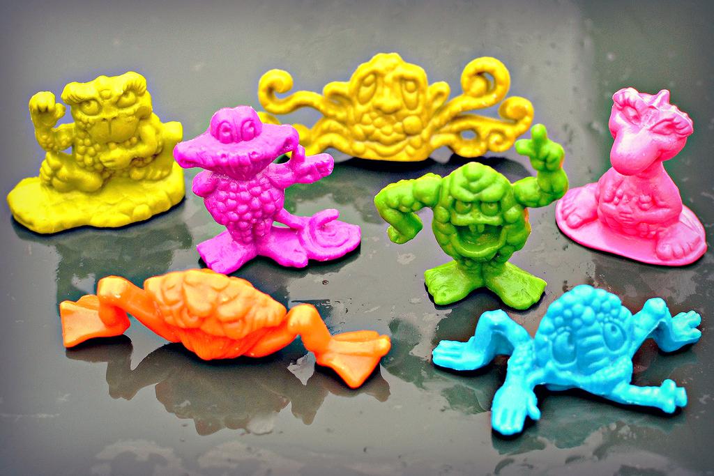Seven brightly colored, small plastic monster and frog toys in yellow, pink, green, orange, and blue are arranged on a wet, dark surface.