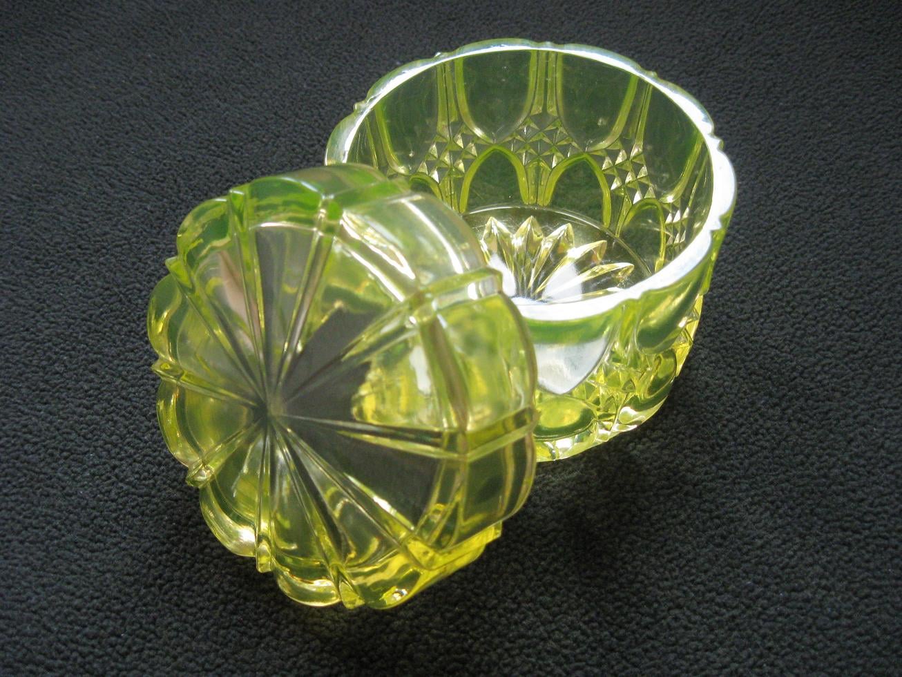 A round, yellow-green glass trinket box with a decorative, faceted pattern and a matching lid resting beside it on a dark textured surface.