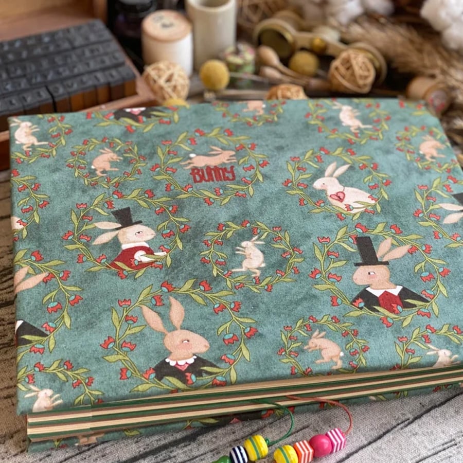 A fabric with a green background decorated with whimsical rabbits, some in suits and top hats, surrounded by leafy wreaths and red flowers. The word “BUNNY” appears among the patterns. Craft supplies are in the background.