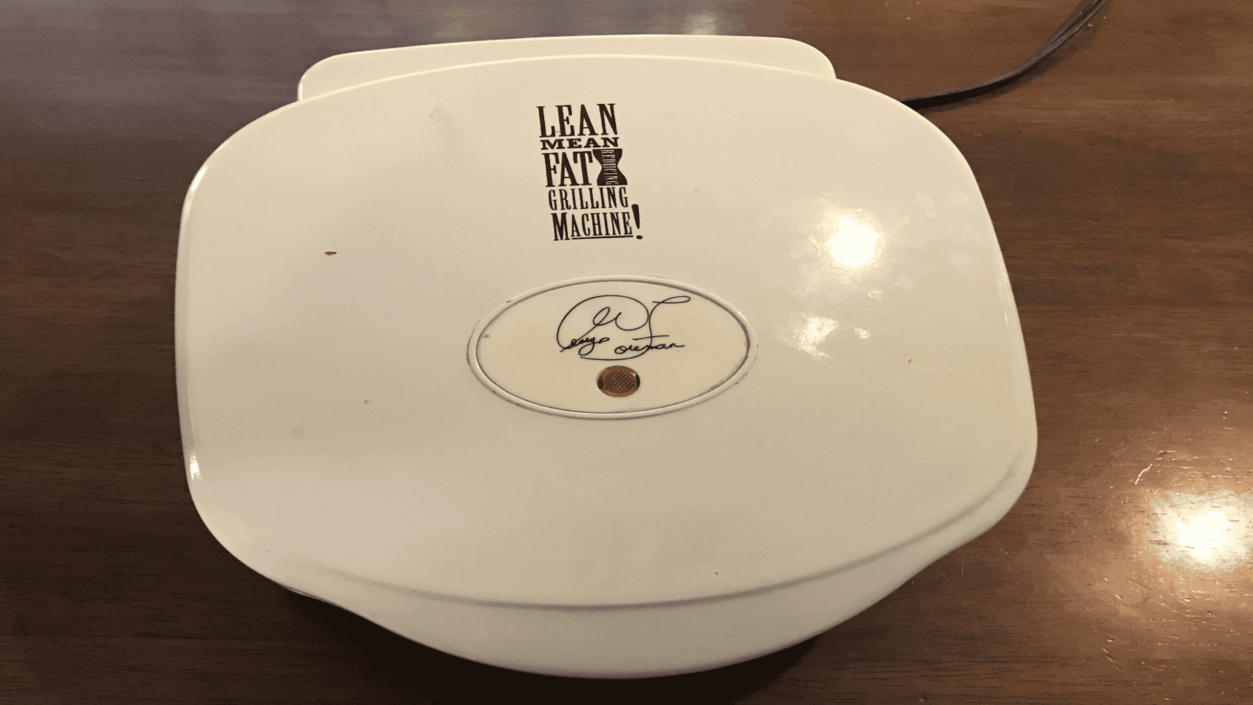 A white George Foreman Grill with the words “Lean Mean Fat-Reducing Grilling Machine!” printed on top, sitting on a wooden table.