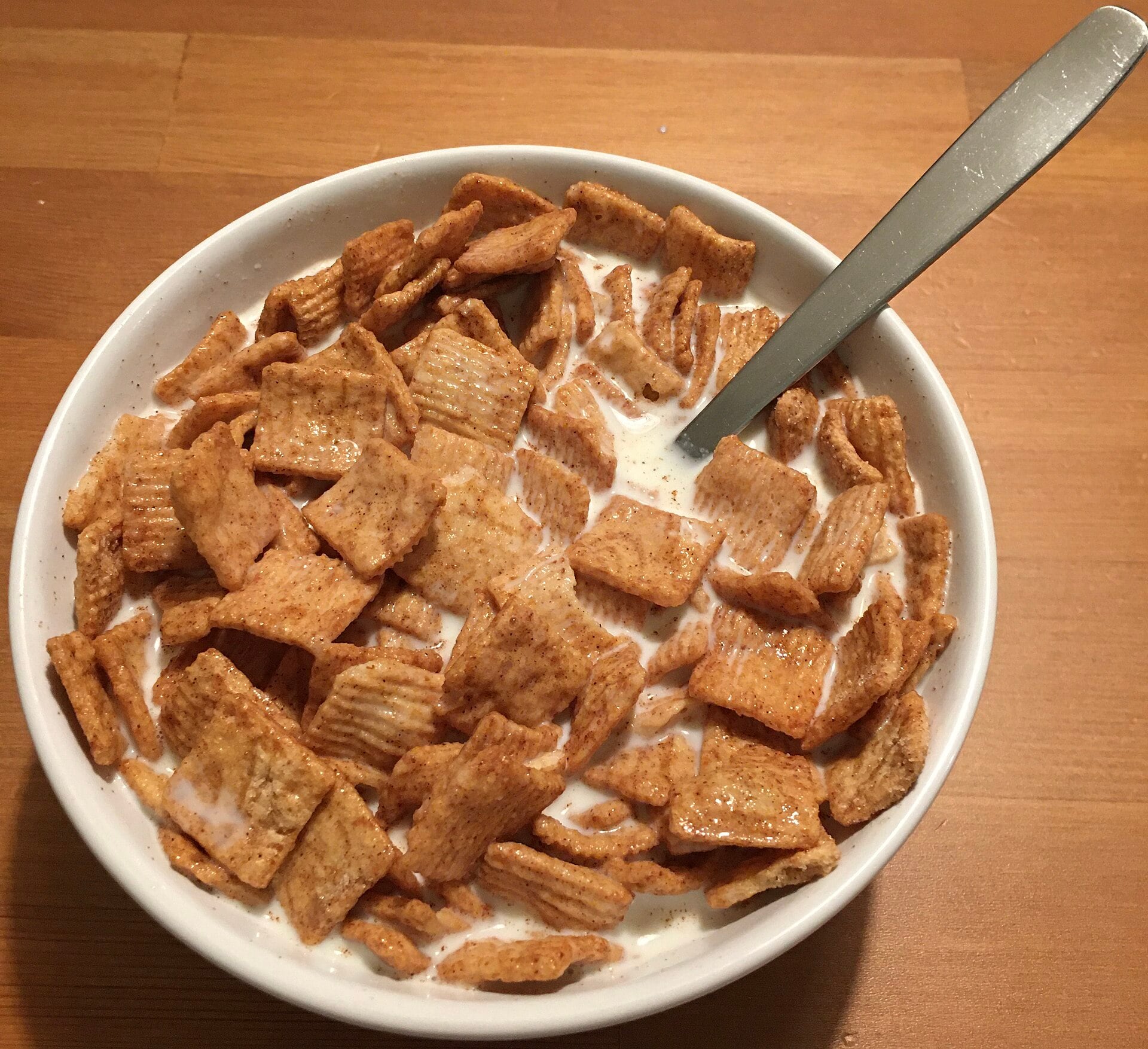 A white bowl filled with cinnamon-flavored square cereal and milk, with a metal spoon resting in the bowl, placed on a wooden table.