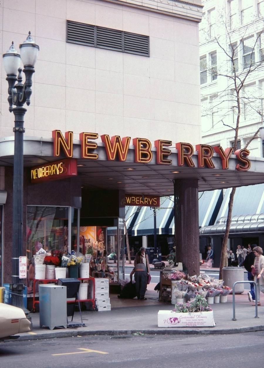 A street view of a Newberrys store entrance with neon signage, outdoor flower stands, and pedestrians walking by. A streetlamp is visible, along with a mix of buildings and trees in the background.