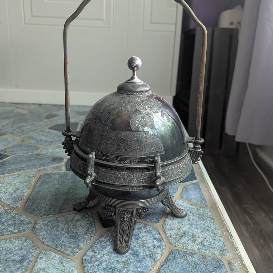 An ornate, vintage metal container with a dome lid, intricate engravings, and four decorative legs stands on a blue-tiled floor near a white wall and purple curtain.