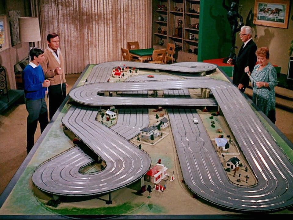Four people stand around a large slot car racetrack set up in a living room, with miniature houses and trees decorating the course. The track has several loops and curves.