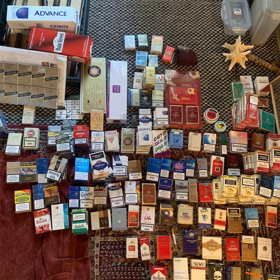 Over 100 cigarette packs of various brands and colors are neatly arranged on a patterned rug and carpet, with some boxes and containers visible around the display.