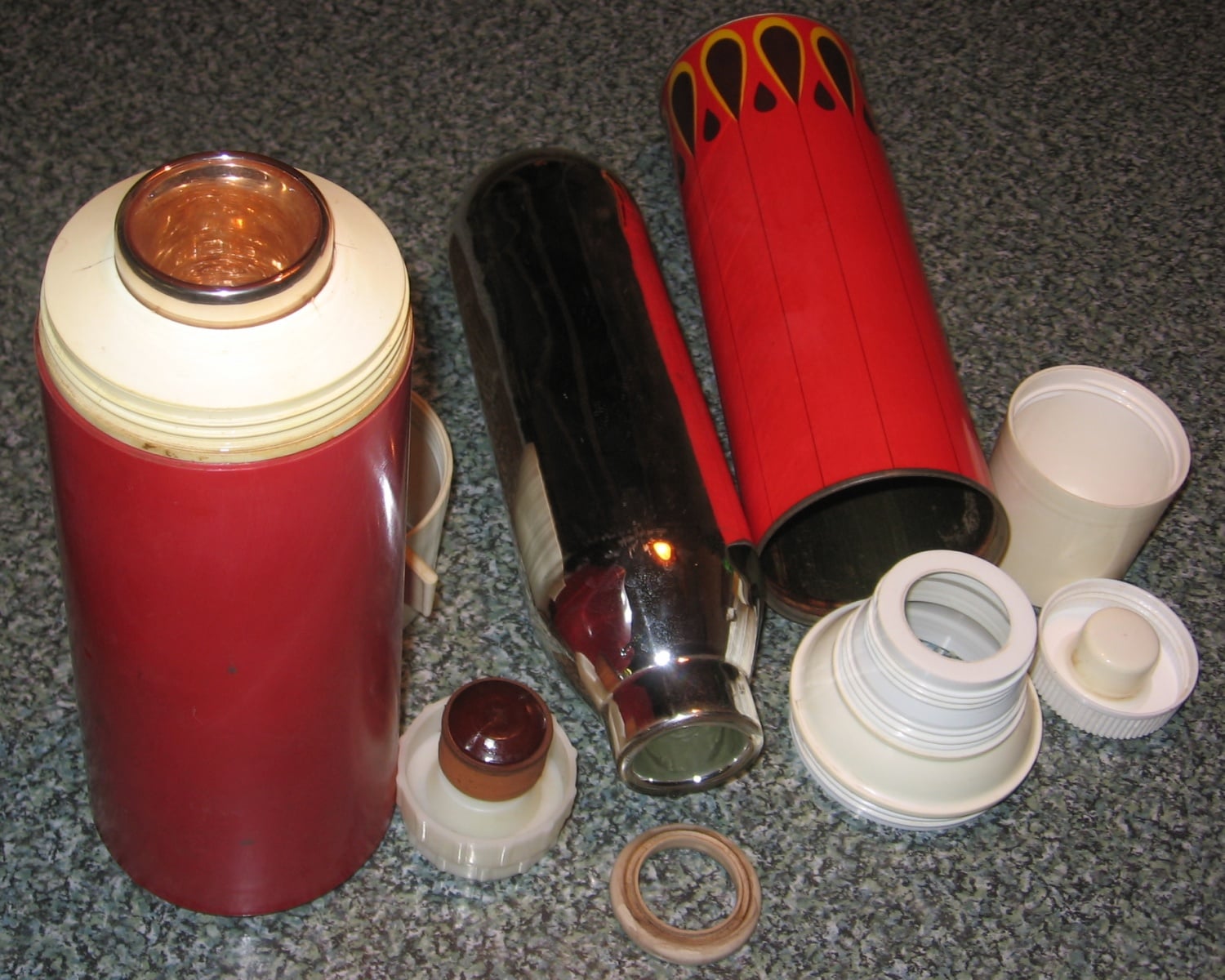 Disassembled red thermos flask showing its metal interior, plastic outer case, lids, and other components, all laid out on a speckled gray surface.