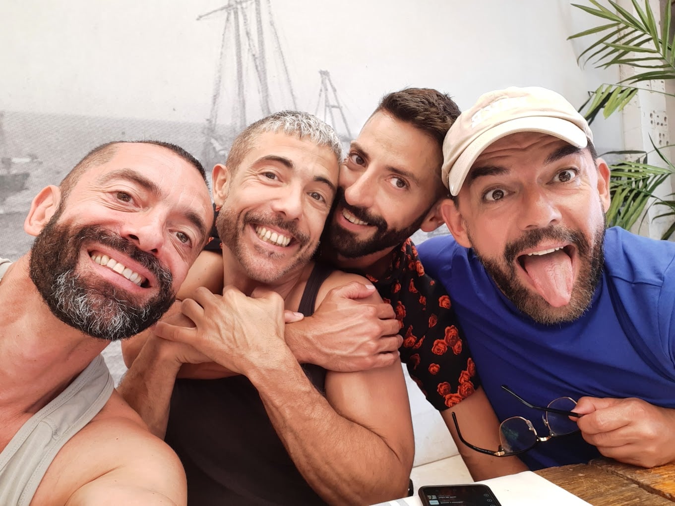 Four smiling men pose for a cheerful selfie; one hugs another from behind, and one sticks out his tongue playfully. They are sitting together indoors near a wall with a sailboat drawing.