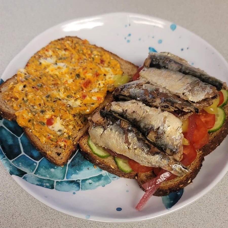 Two slices of whole grain bread: one topped with a spread and herbs, the other with cucumber, tomato, red onion, and several sardines, served on a white plate with a blue pattern.