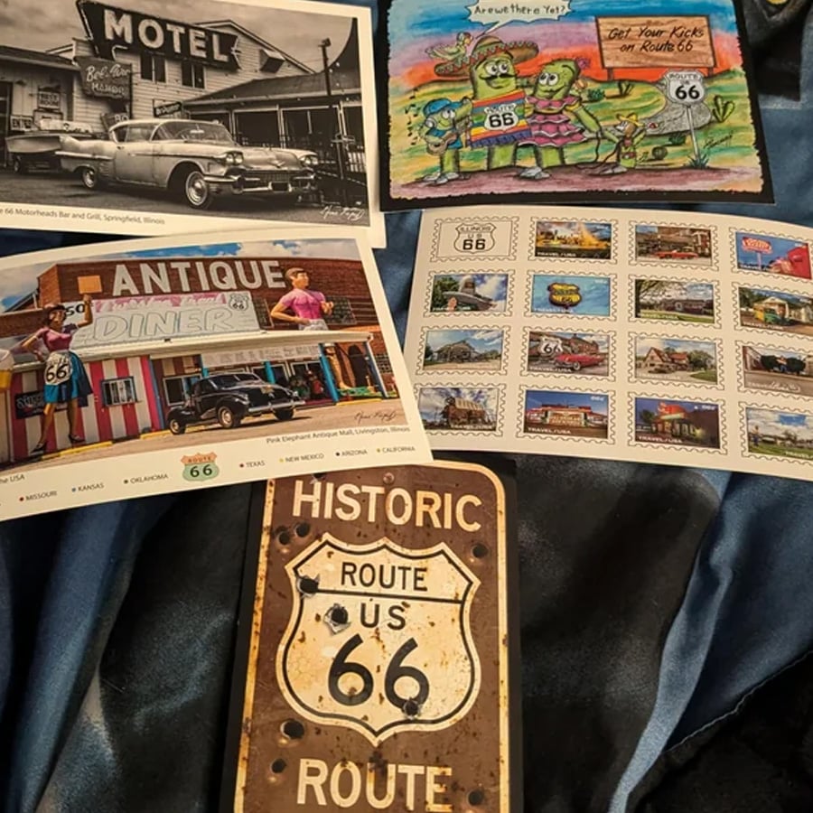 Postcards, stamps, and a metal sign featuring Route 66 memorabilia and artwork are arranged on a dark cloth, highlighting classic cars, diners, and cartoon characters themed around the historic American highway.