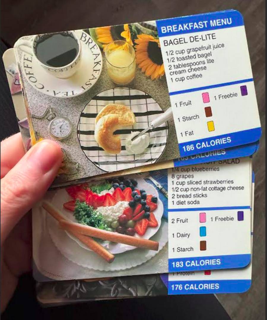 A hand holds meal cards showing plated breakfasts with calorie counts, food group breakdowns, and menu details like bagel, fruit, and coffee. The cards are color-coded and display meal options and nutritional information.