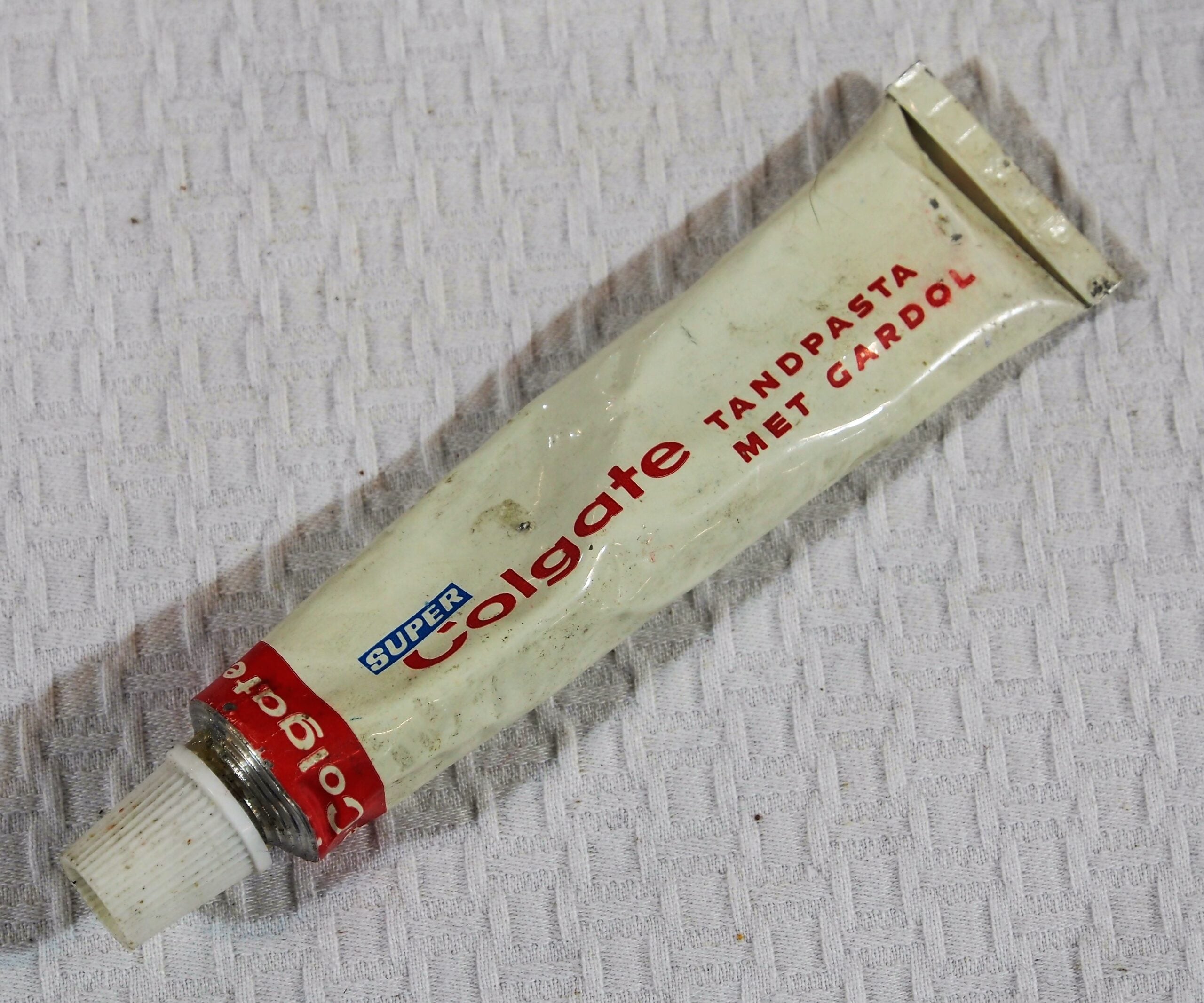 Old, worn tube of “Super Colgate Tandpasta Met Gardol” toothpaste with a white cap, lying on a textured white surface. The tube is slightly bent and discolored.