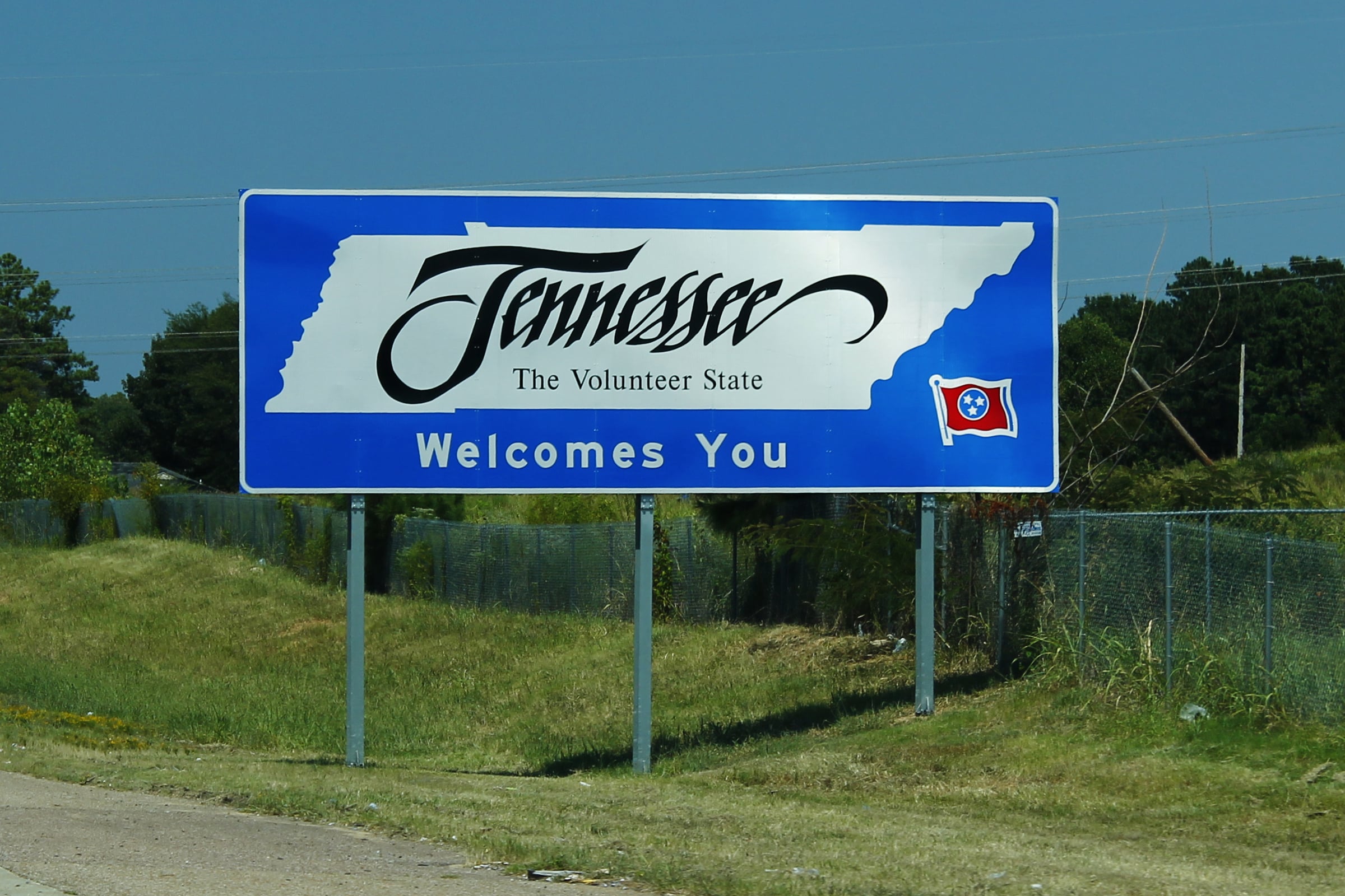 A blue and white roadside sign reads “Tennessee The Volunteer State Welcomes You,” with the state outline and flag on it, beside a grassy area and fence.