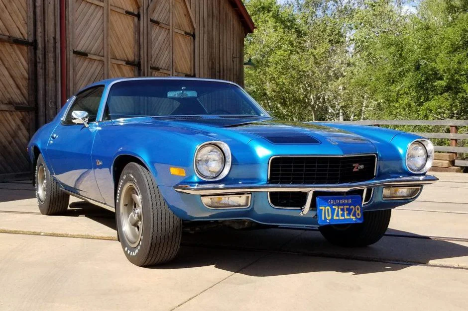 A blue classic Chevrolet Camaro Z28 parked on a driveway in front of a wooden barn, with trees and sunlight in the background.